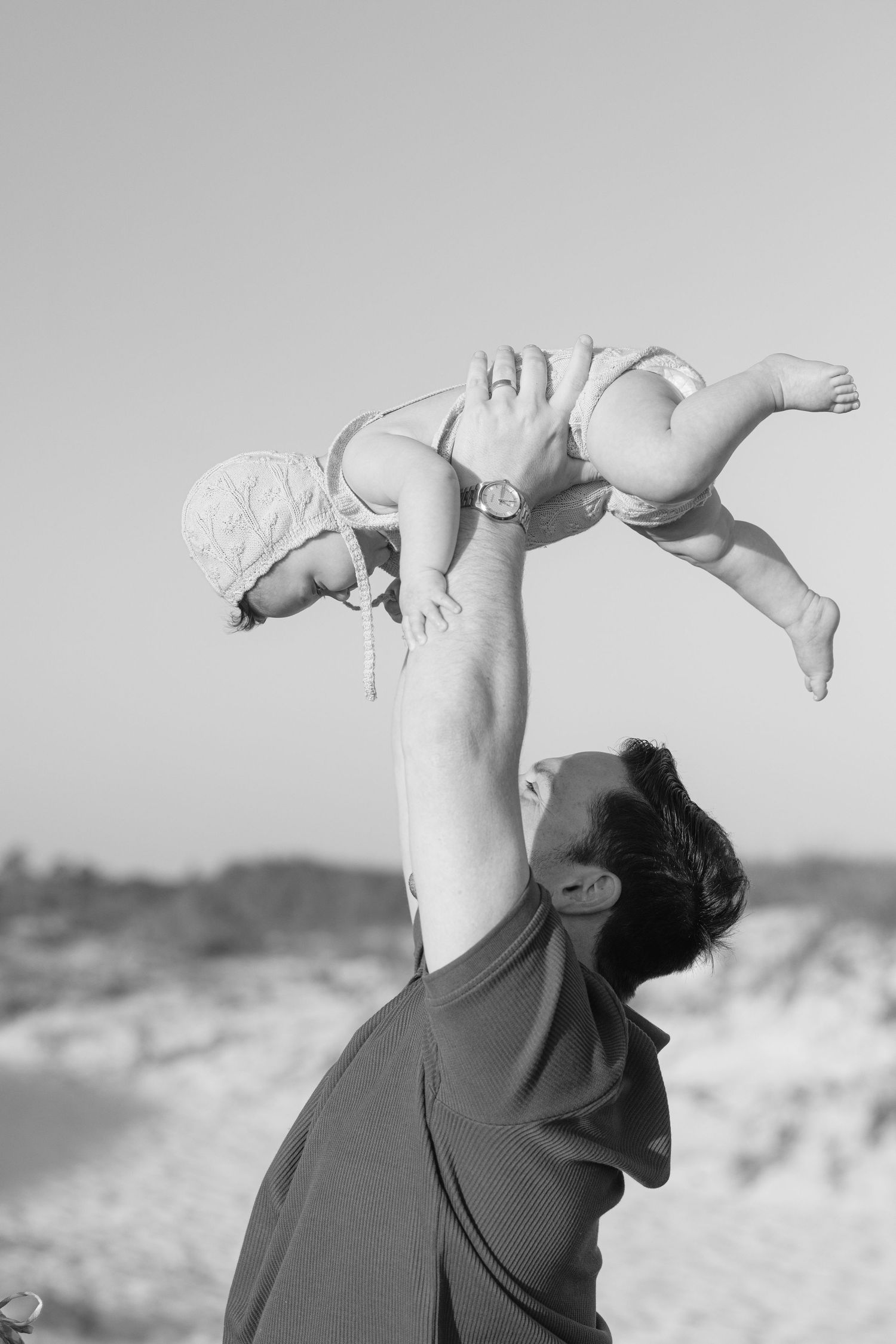 Black and white photo of a parent lifting a baby overhead at the beach, sharing a joyful moment outdoors.