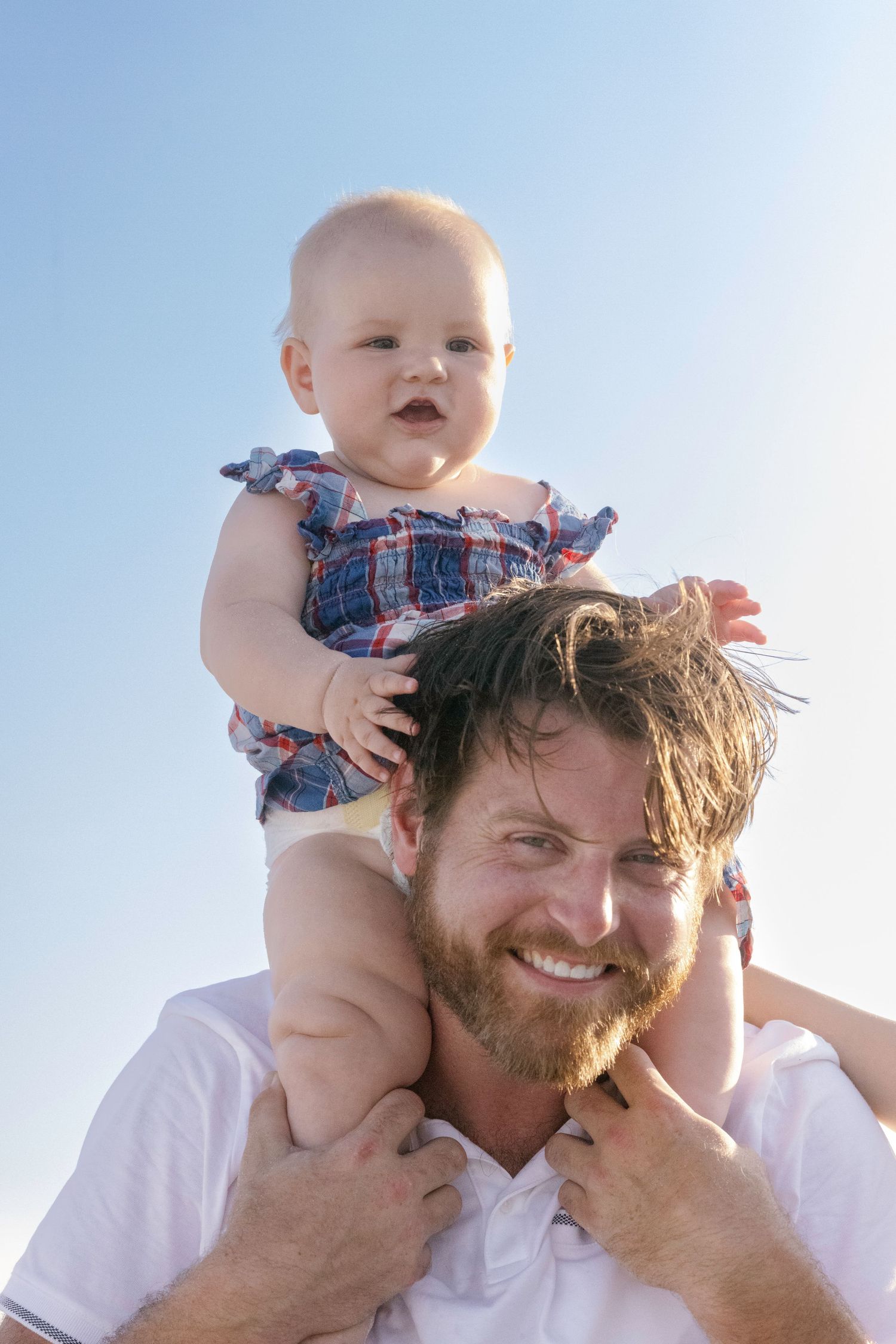 Smiling bearded father carrying happy baby girl on his shoulders against a clear blue sky.