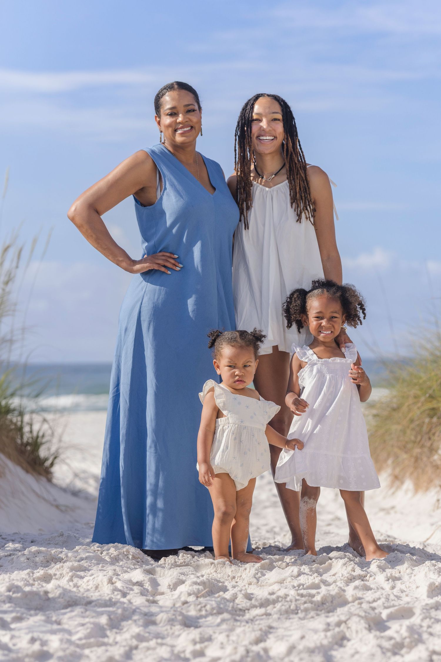 Happy African American family of four, two women and two little girls in white dresses, posing on a sandy beach.