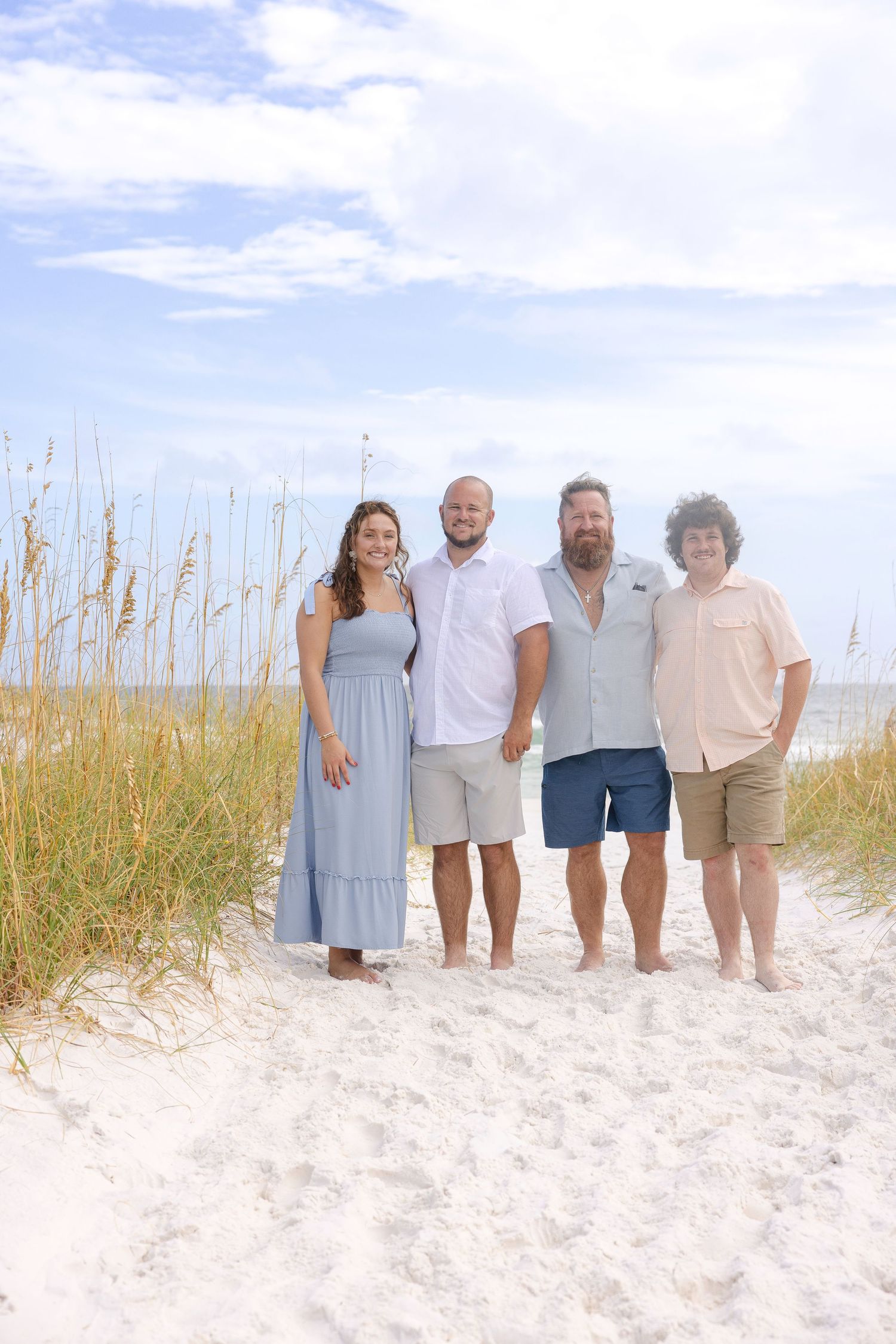 Family group standing together on white sandy beach with sea oats and blue sky in the background.