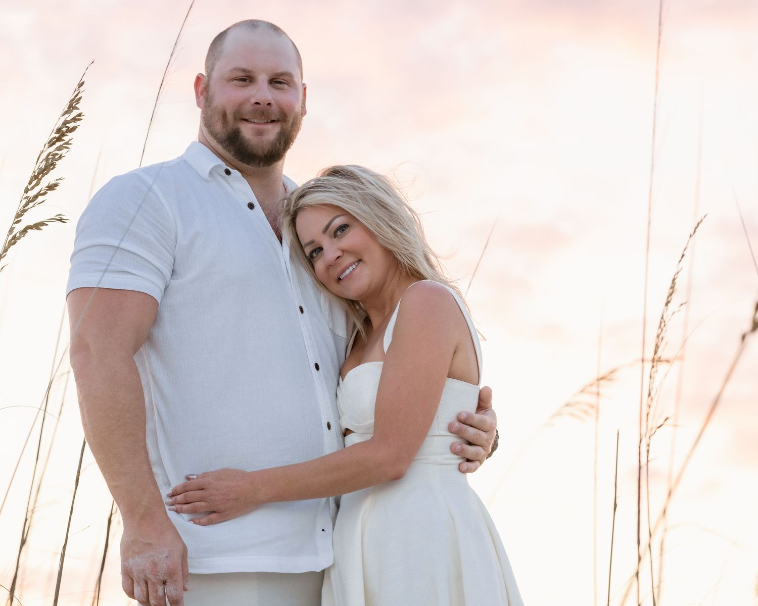 Smiling couple in white outfits embrace among sea oats at a colorful beach sunset.