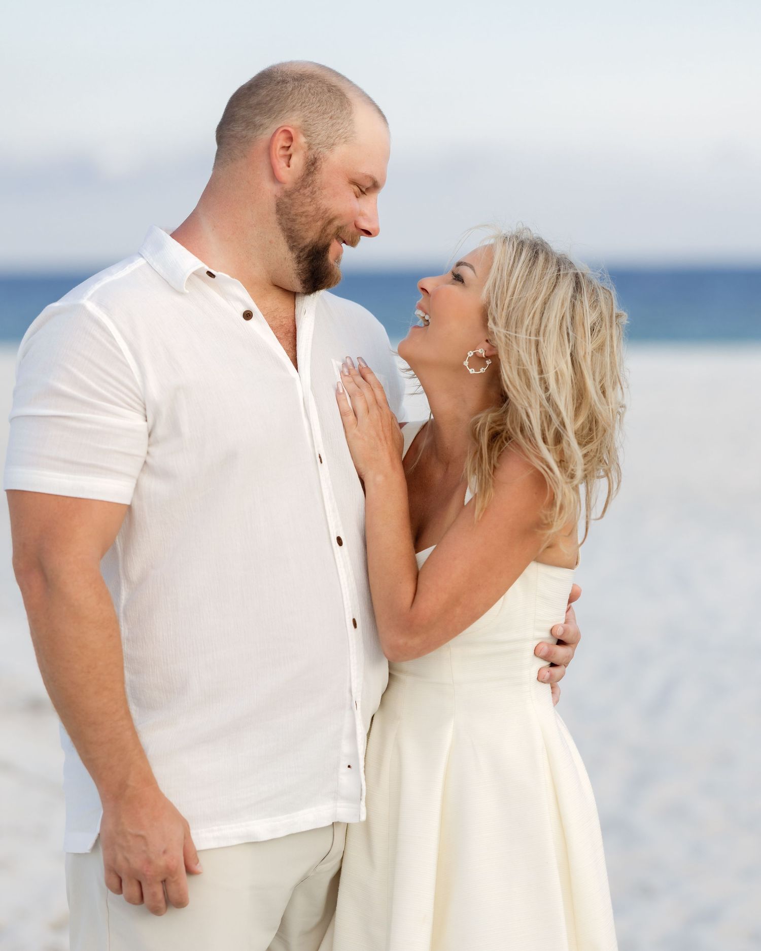 Happy couple in matching white outfits embrace and smile at each other on a sunny beach during a photo session.
