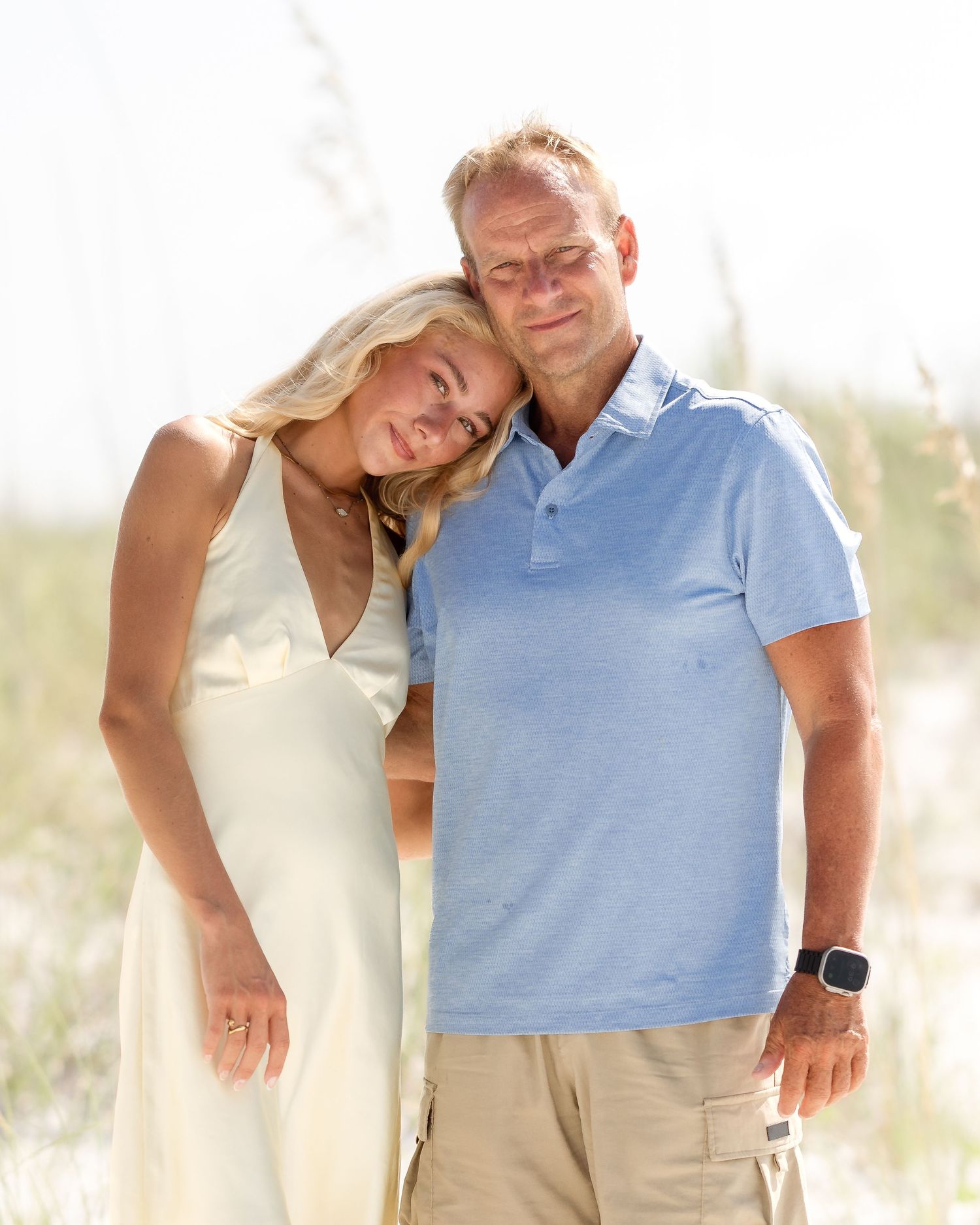 A couple poses together on a sandy beach, woman in white dress leaning on mans shoulder, wearing blue polo shirt.