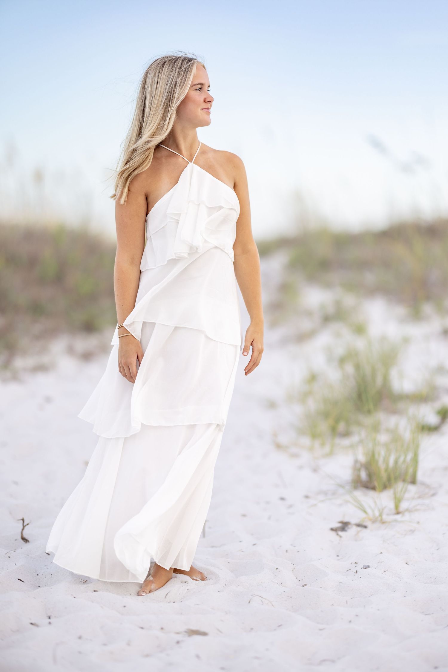 Woman in white tiered halter maxi dress standing on sandy beach dunes with sea oats in background.