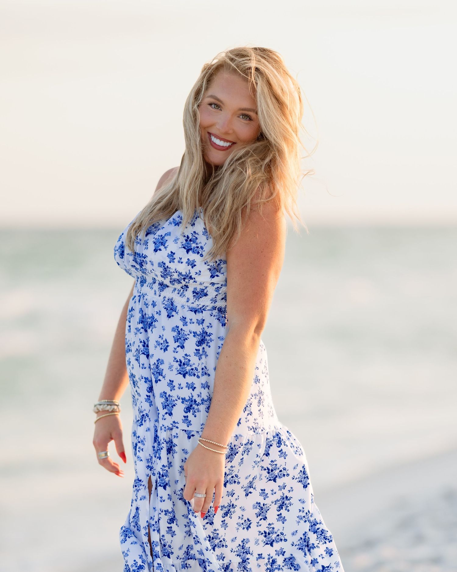 Smiling blonde woman in blue floral dress laughing on sunny beach with ocean backdrop.