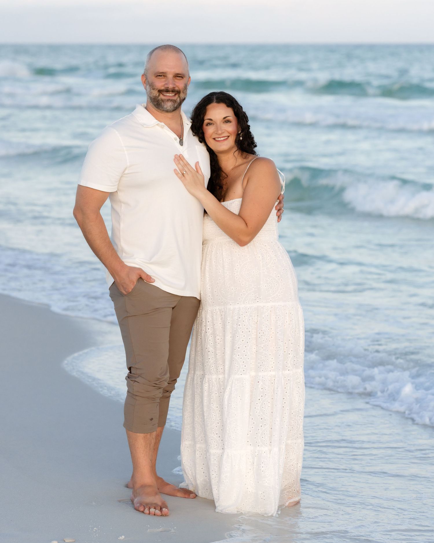 Couple posing together on a sandy beach, man in white shirt and woman in white maxi dress, with ocean waves behind them.