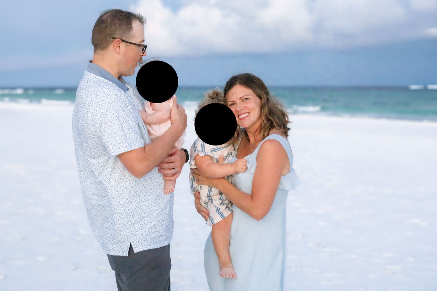 Family of four posing on a sandy beach with turquoise ocean waves and cloudy blue sky in the background.