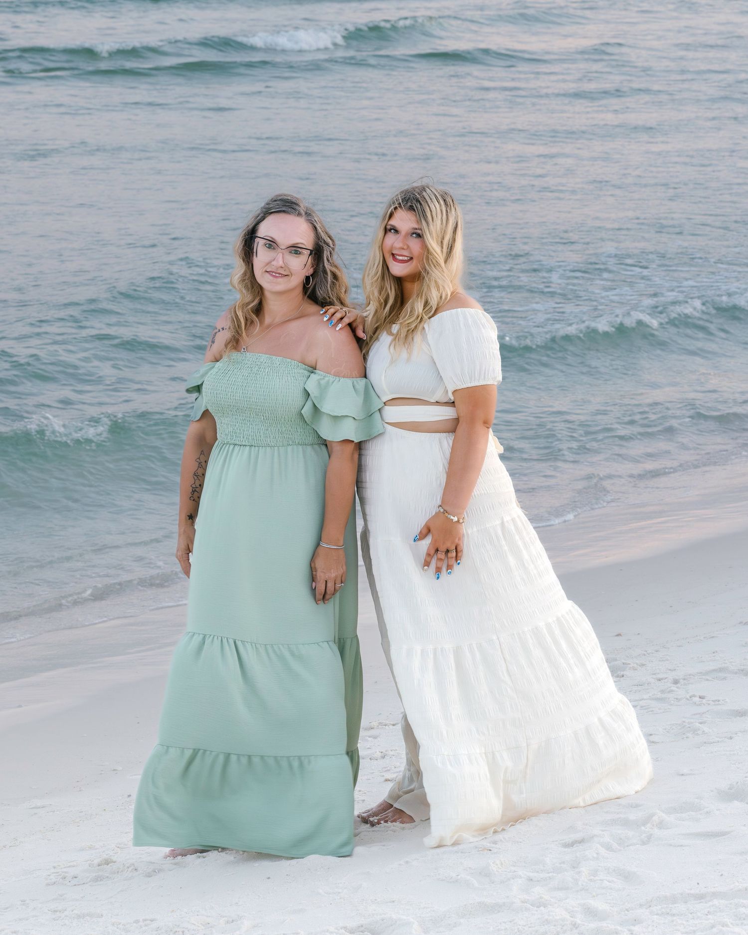 Group sits together on sandy beach with ocean waves in background wearing pastel summer dresses.