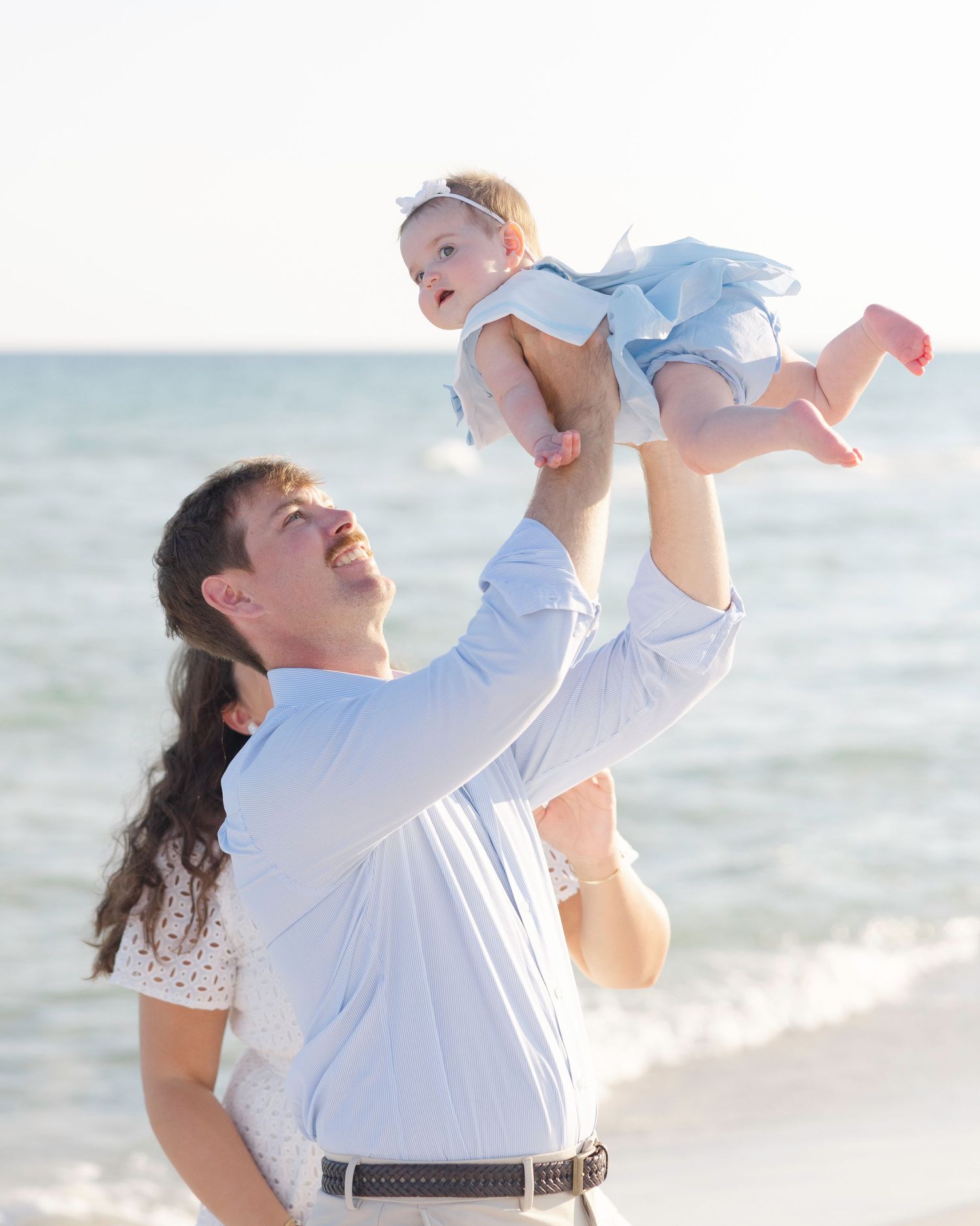 A couple in white outfits plays joyfully with their baby on a sunny beach at sunset.
