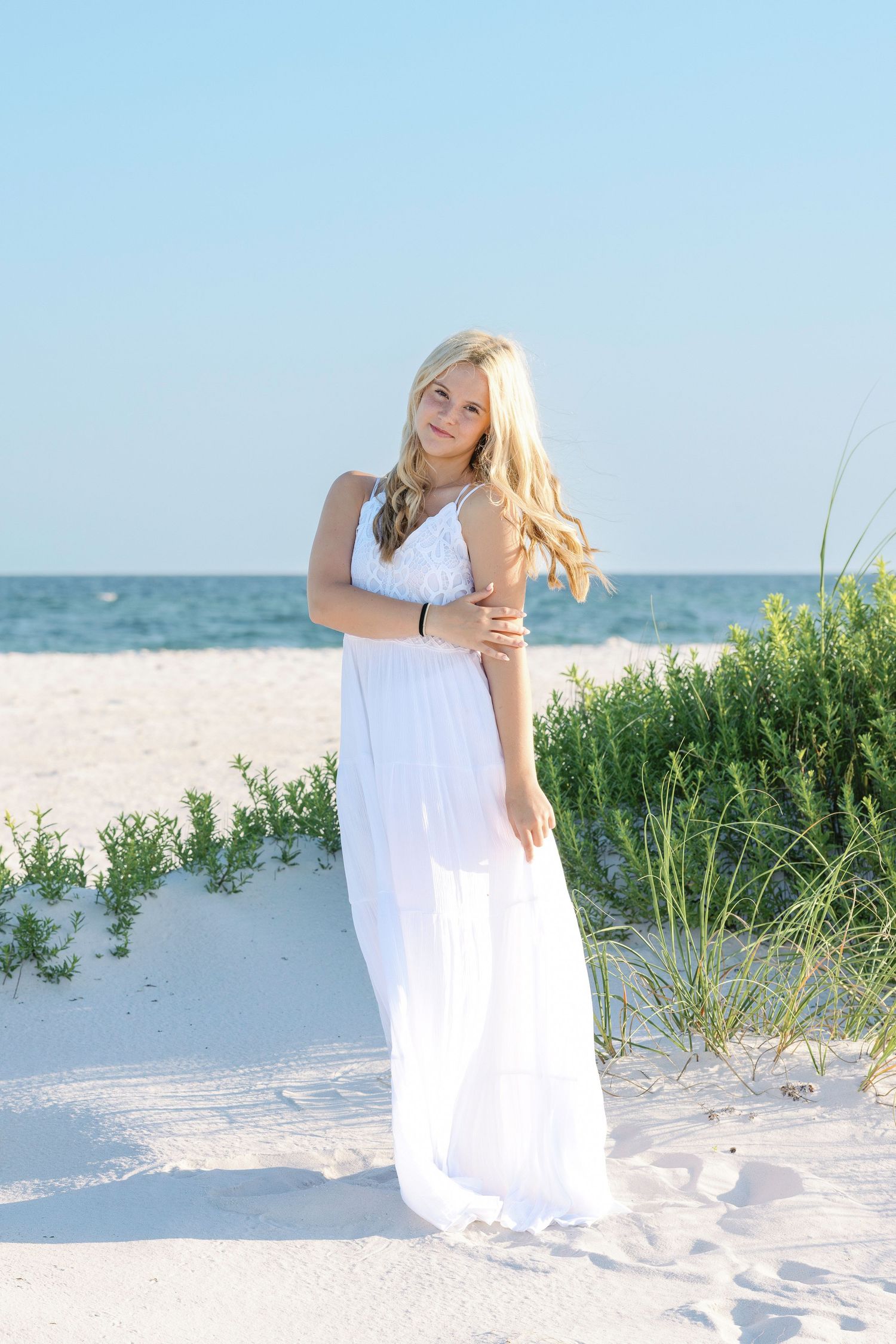 Woman in flowing white maxi dress poses on sandy beach with ocean and dune grass in background.