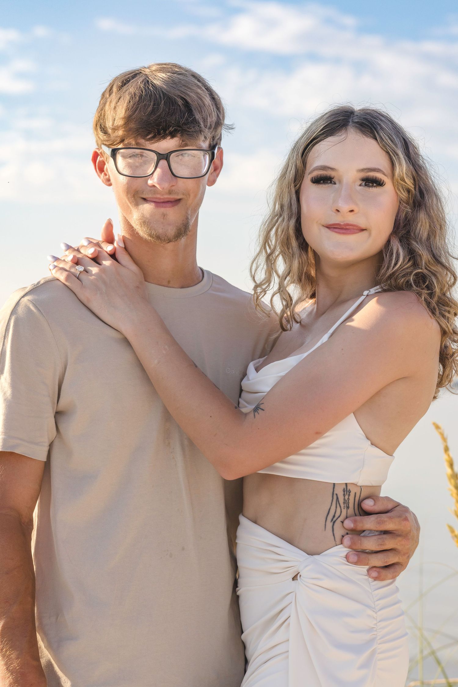 A couple shares intimate moments on a beach at sunset wearing beige and white outfits.