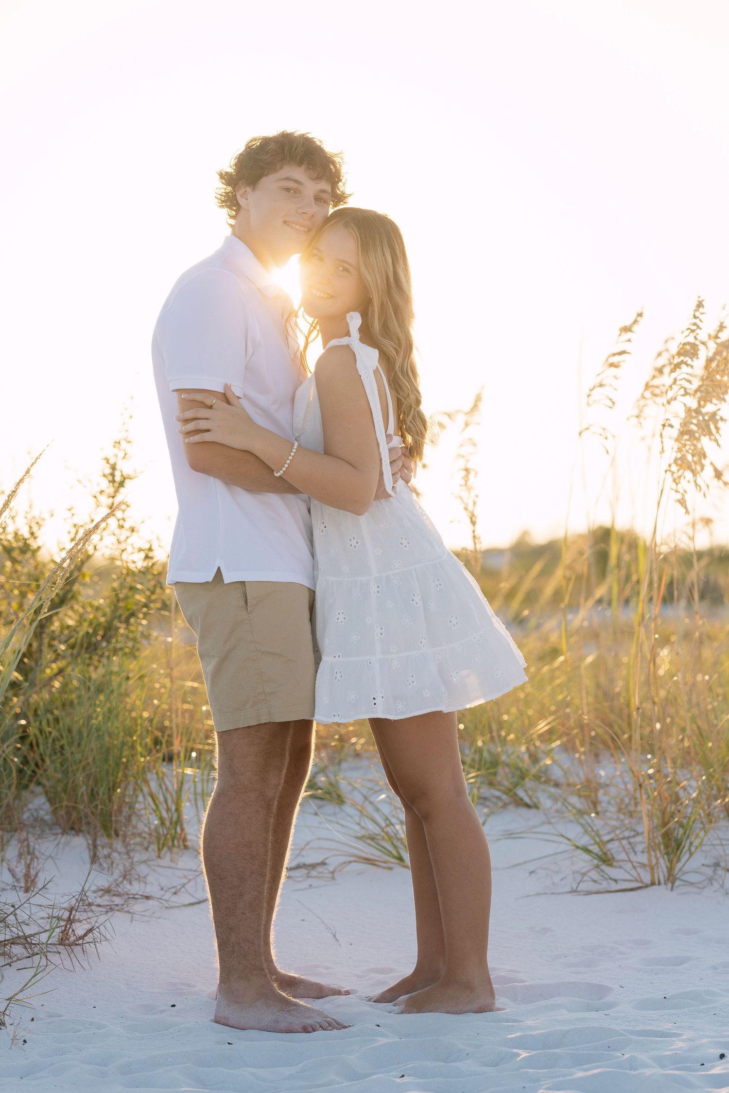 A romantic couple embraces on a sandy beach at sunset with sea oats swaying in the golden light.