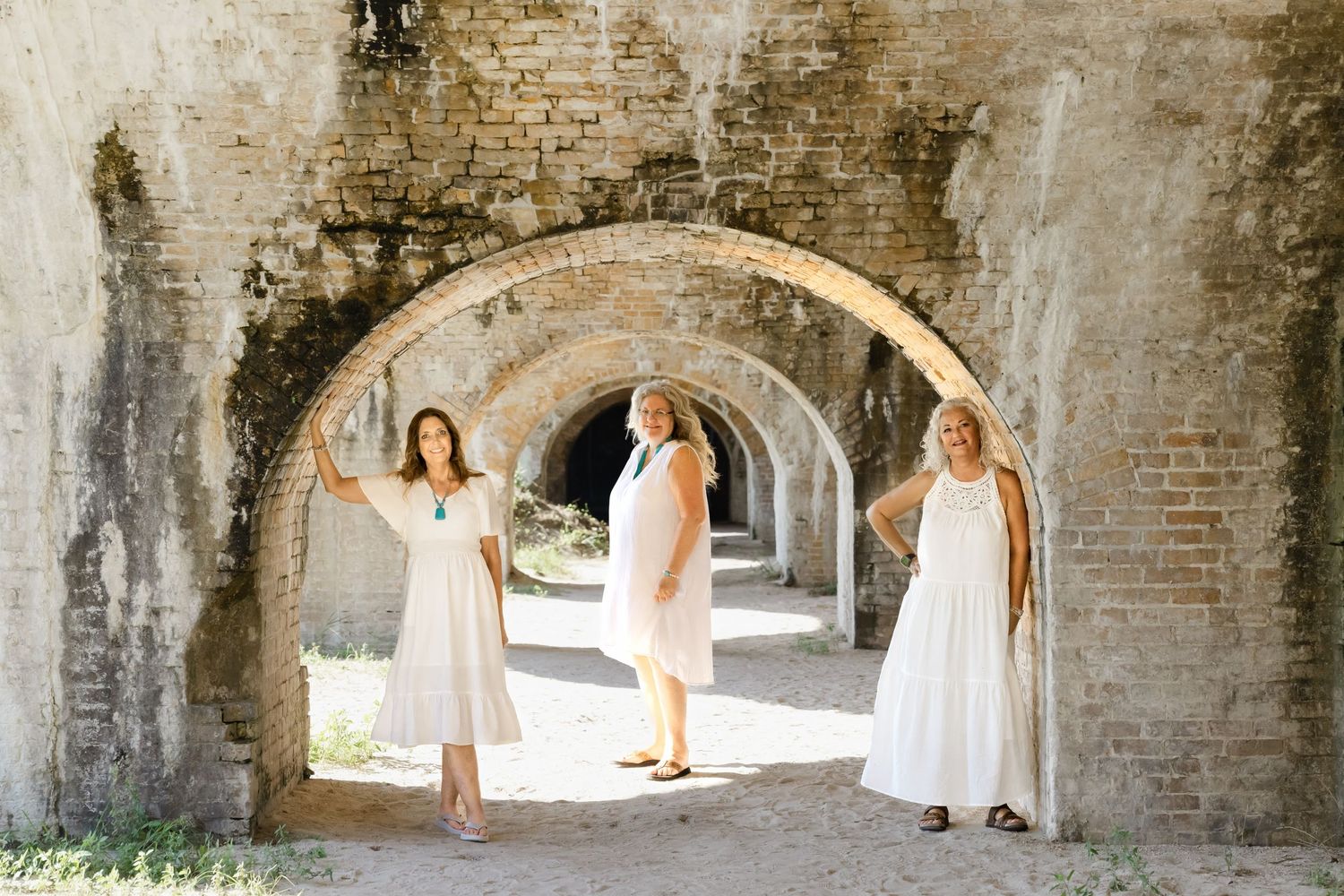 People in white dresses walk through an ancient stone archway with rustic brick walls in soft natural lighting.