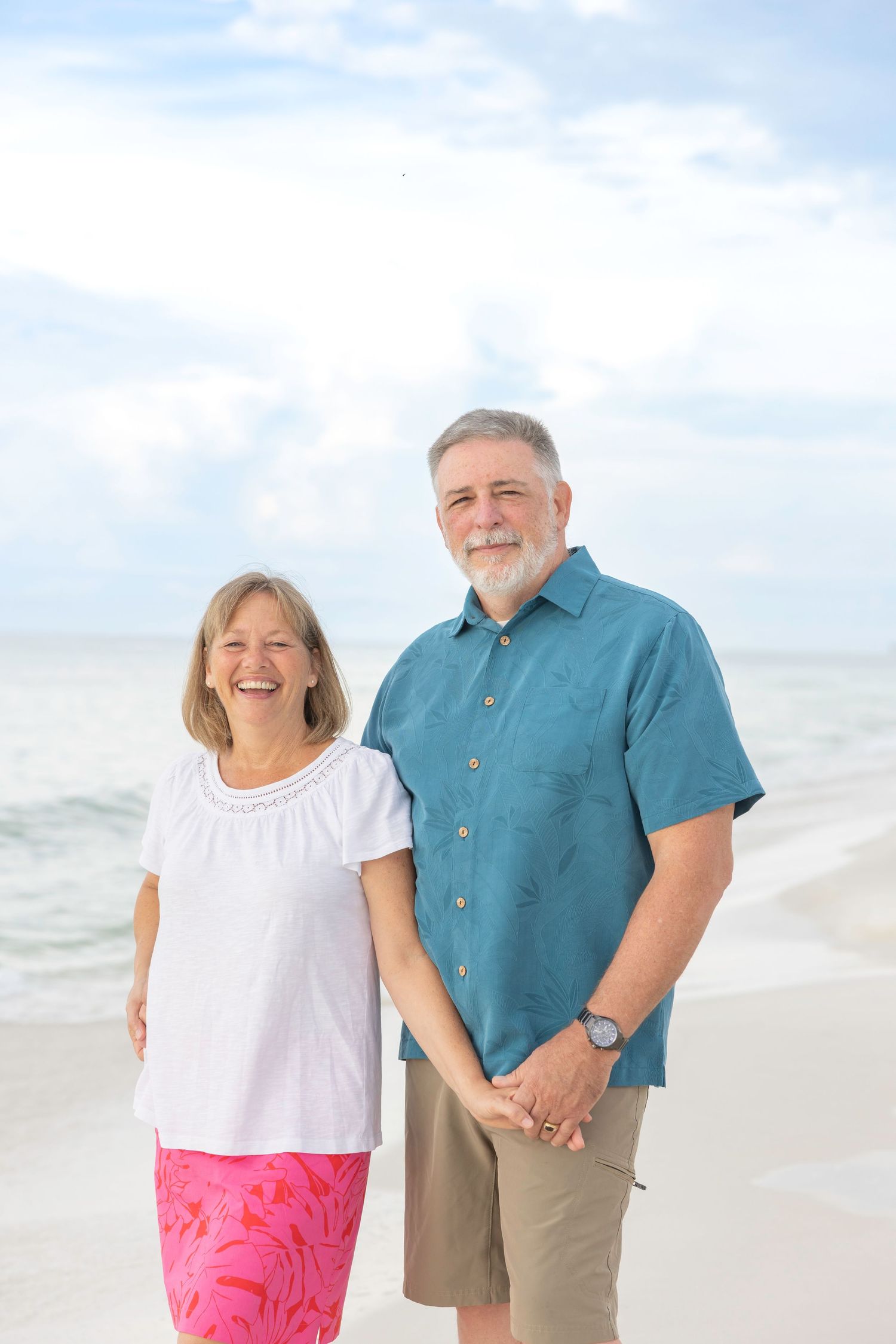Couple poses together on a bright sandy beach with ocean backdrop in casual summer attire.