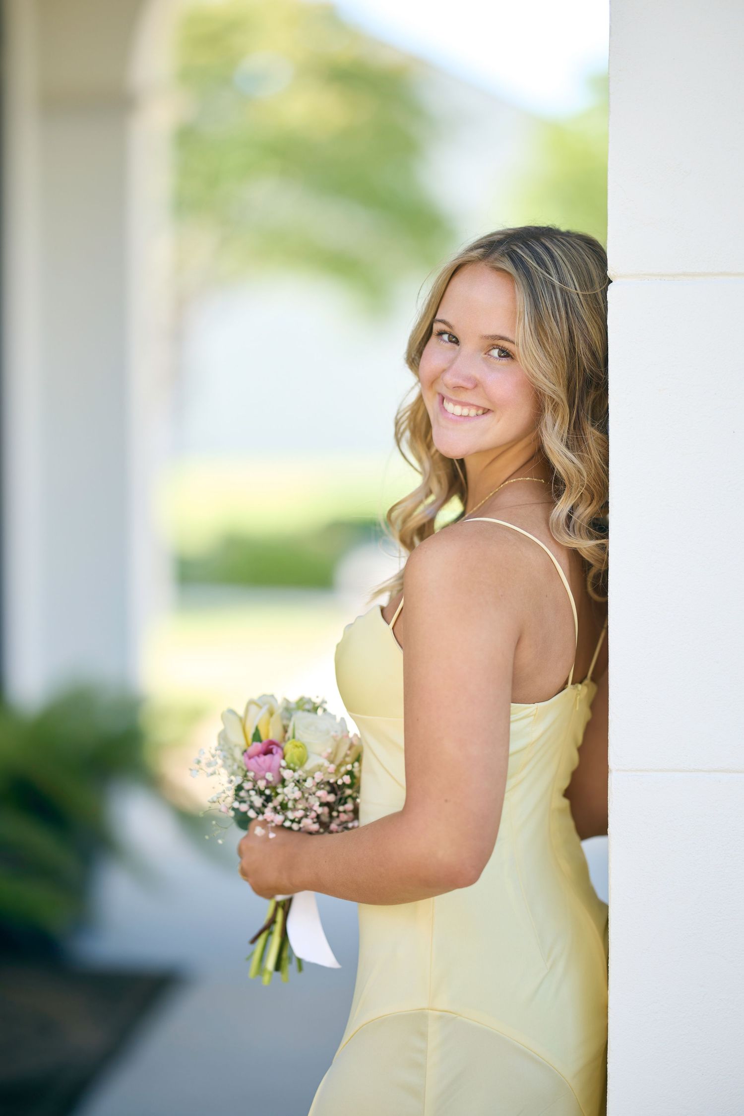 A bridesmaid in a soft yellow gown poses with a bouquet against white columns outdoors.