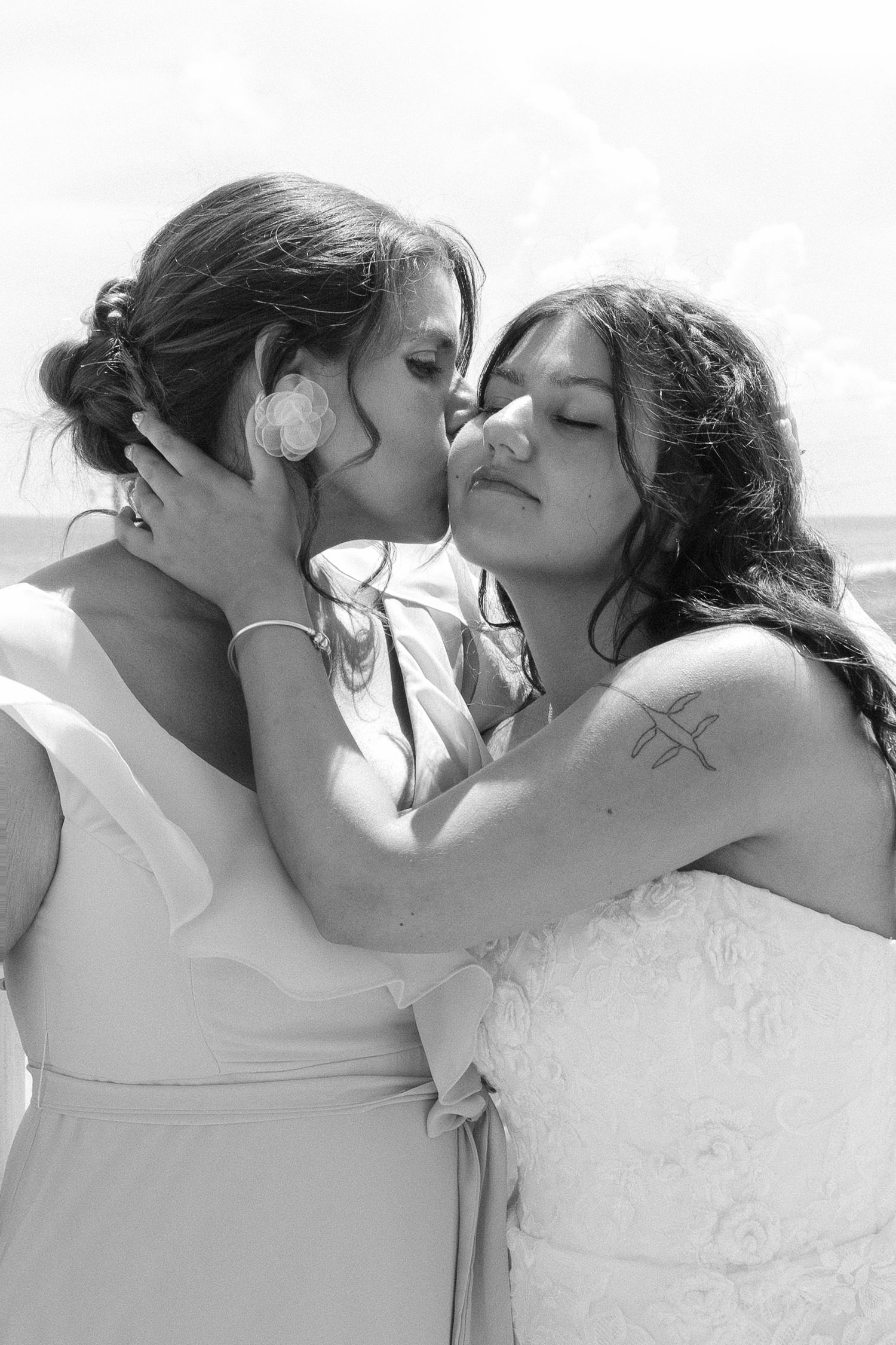 Two people in white dresses share an intimate moment in black and white photography.