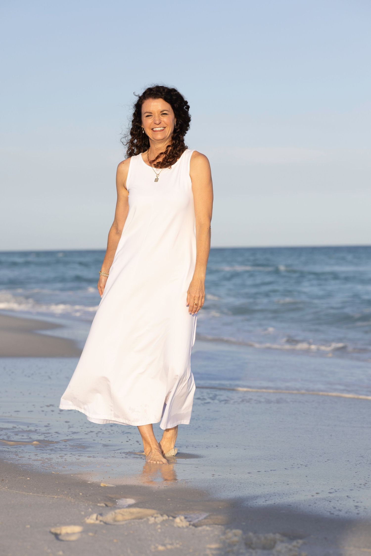 A smiling woman in a white maxi dress walks barefoot along a sunny beach with ocean waves in the background.