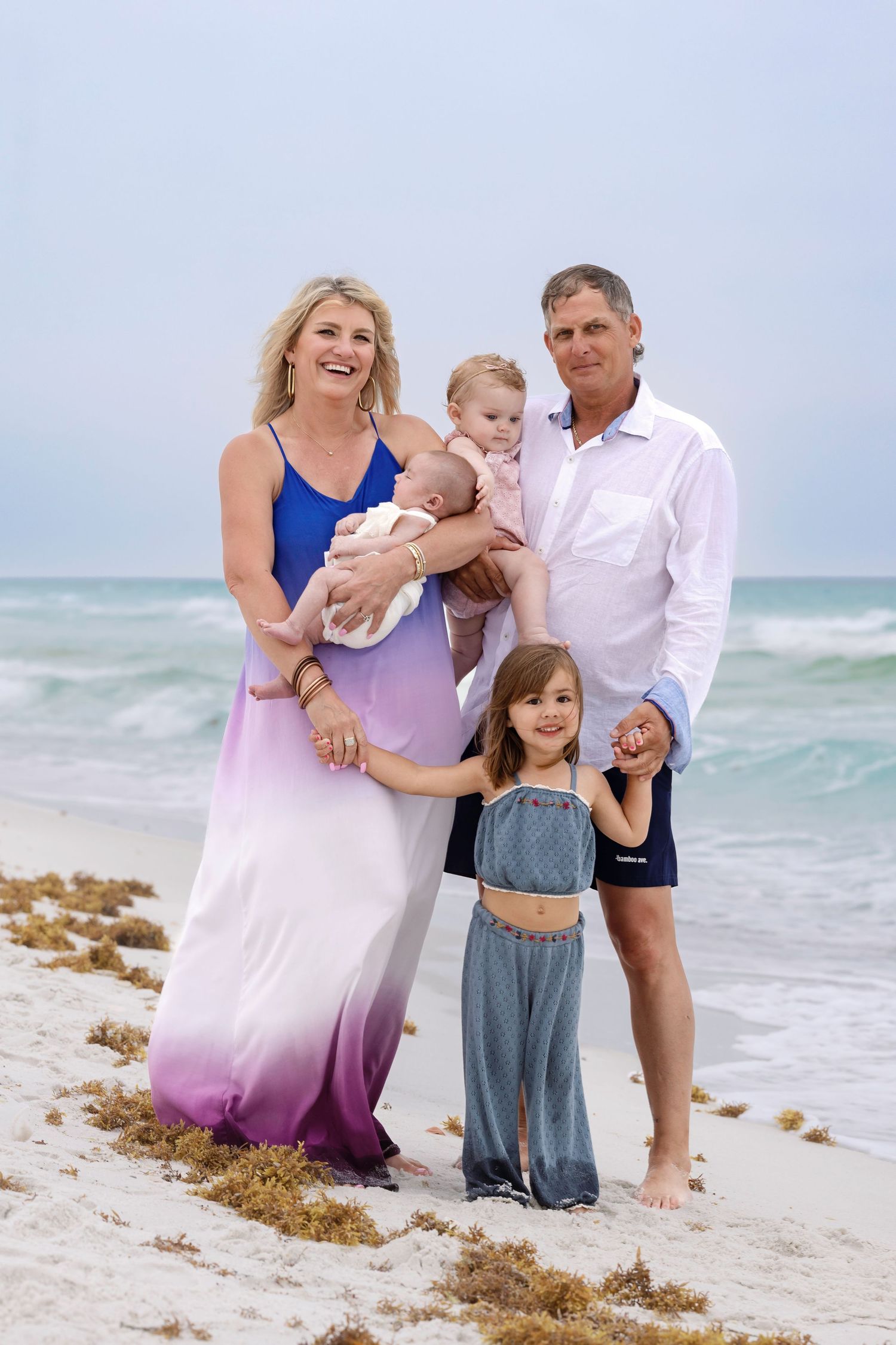 Family of five posing on a sandy beach, with parents holding young children, ocean waves in the background.