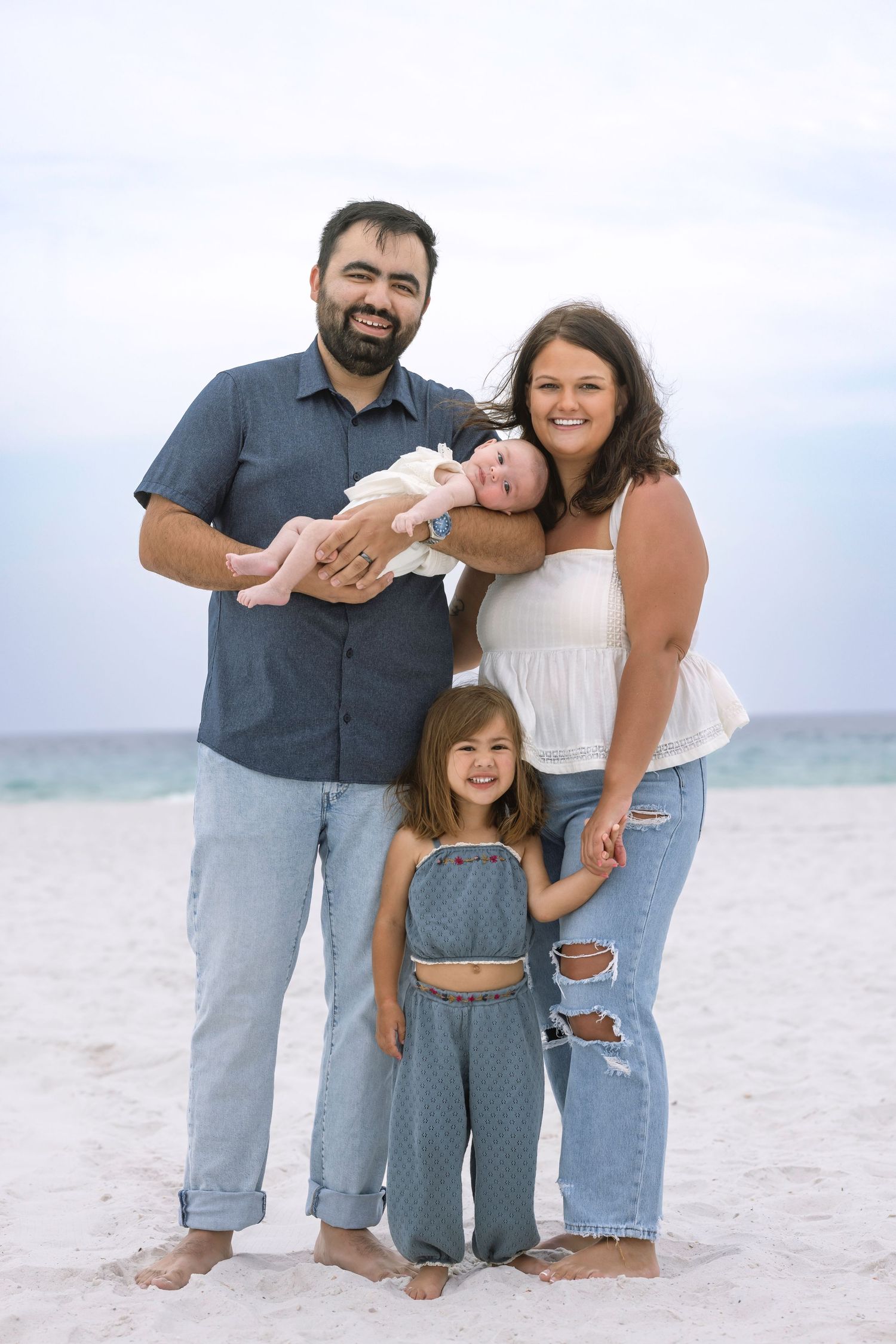 Happy family of four poses on white sand beach, parents holding newborn and young daughter in matching denim outfits.