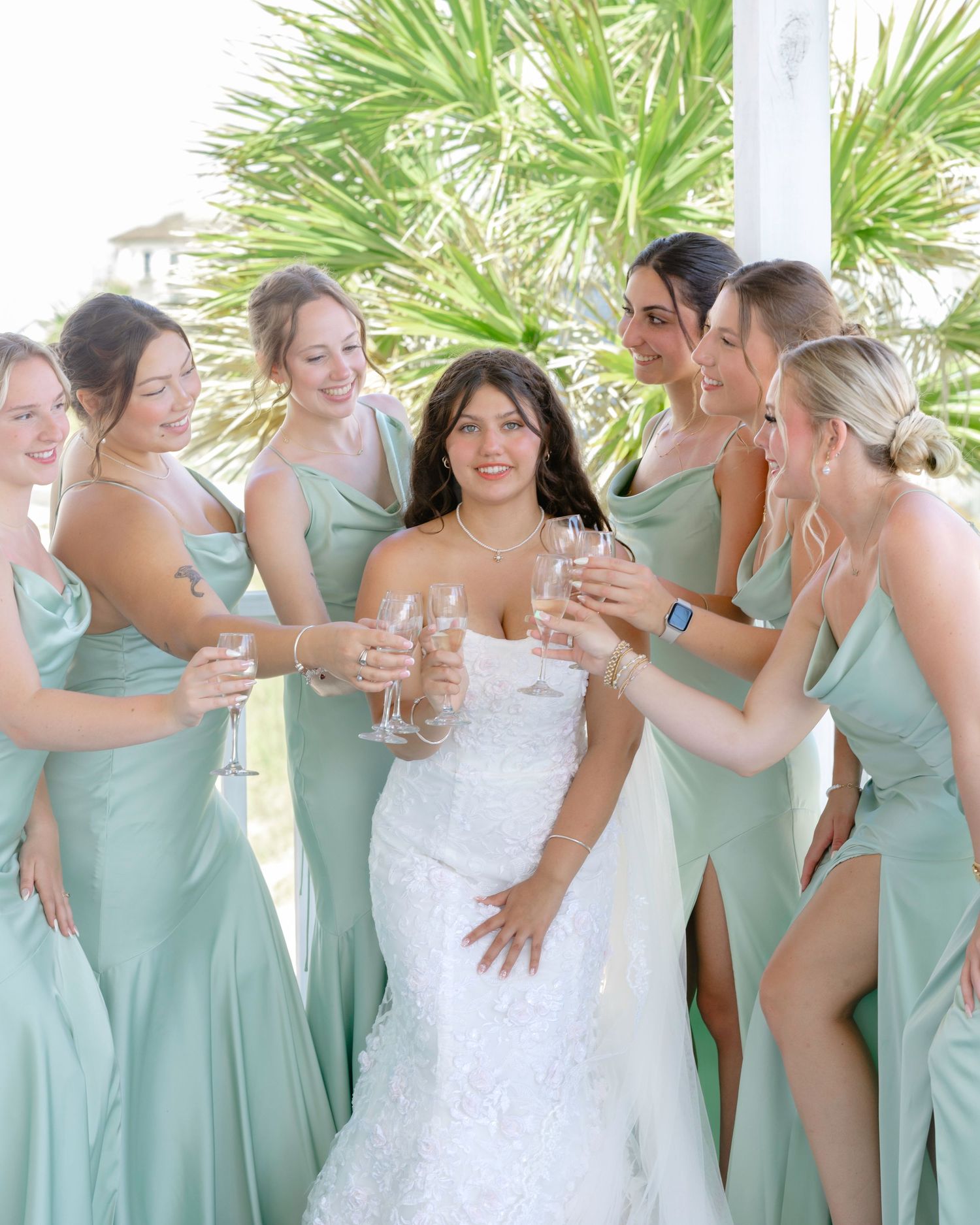 Bridesmaids in sage green gowns toast the bride in a white lace dress outdoors with champagne.