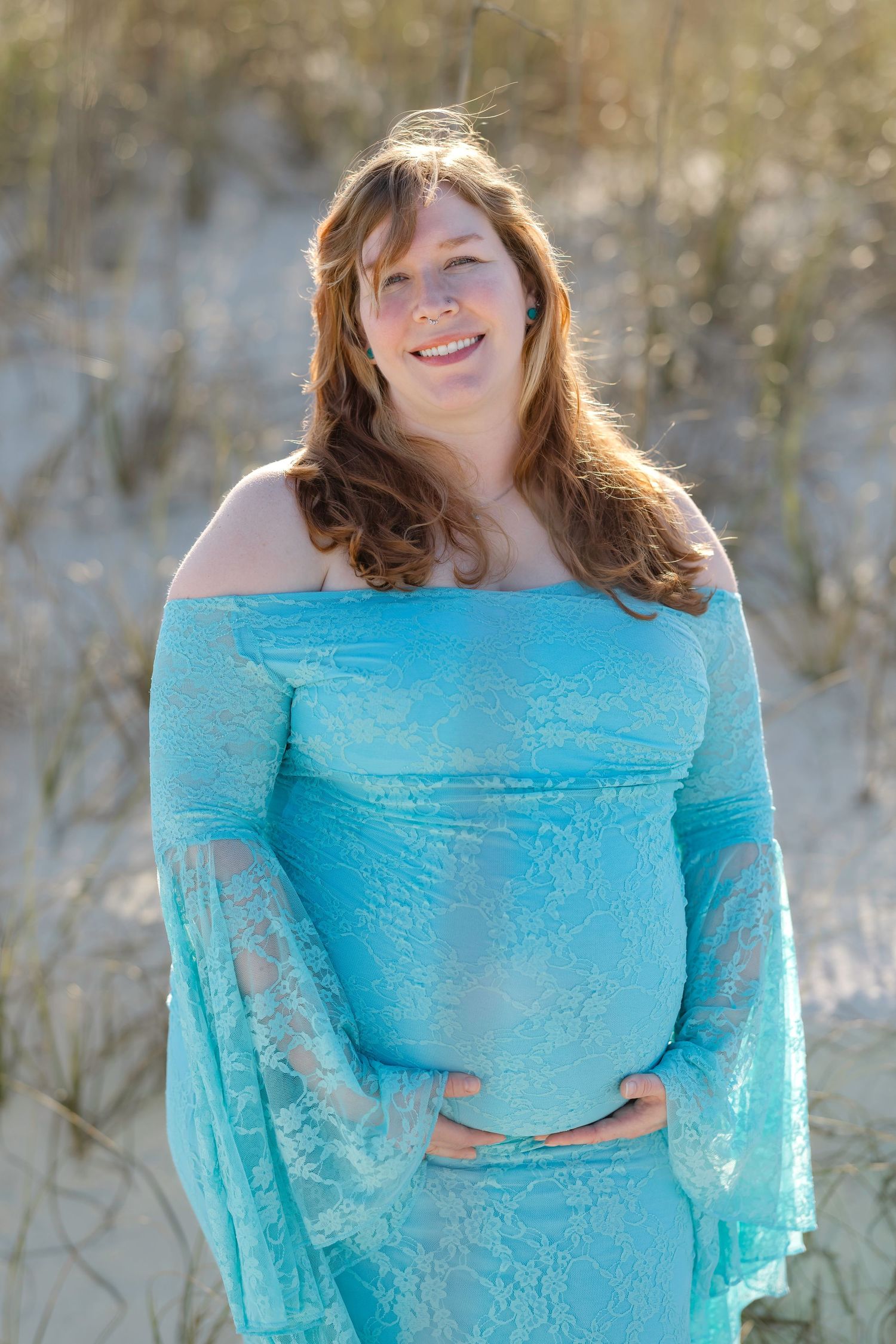 Smiling pregnant woman in turquoise lace off-shoulder maternity gown cradling baby bump on sandy beach dunes.