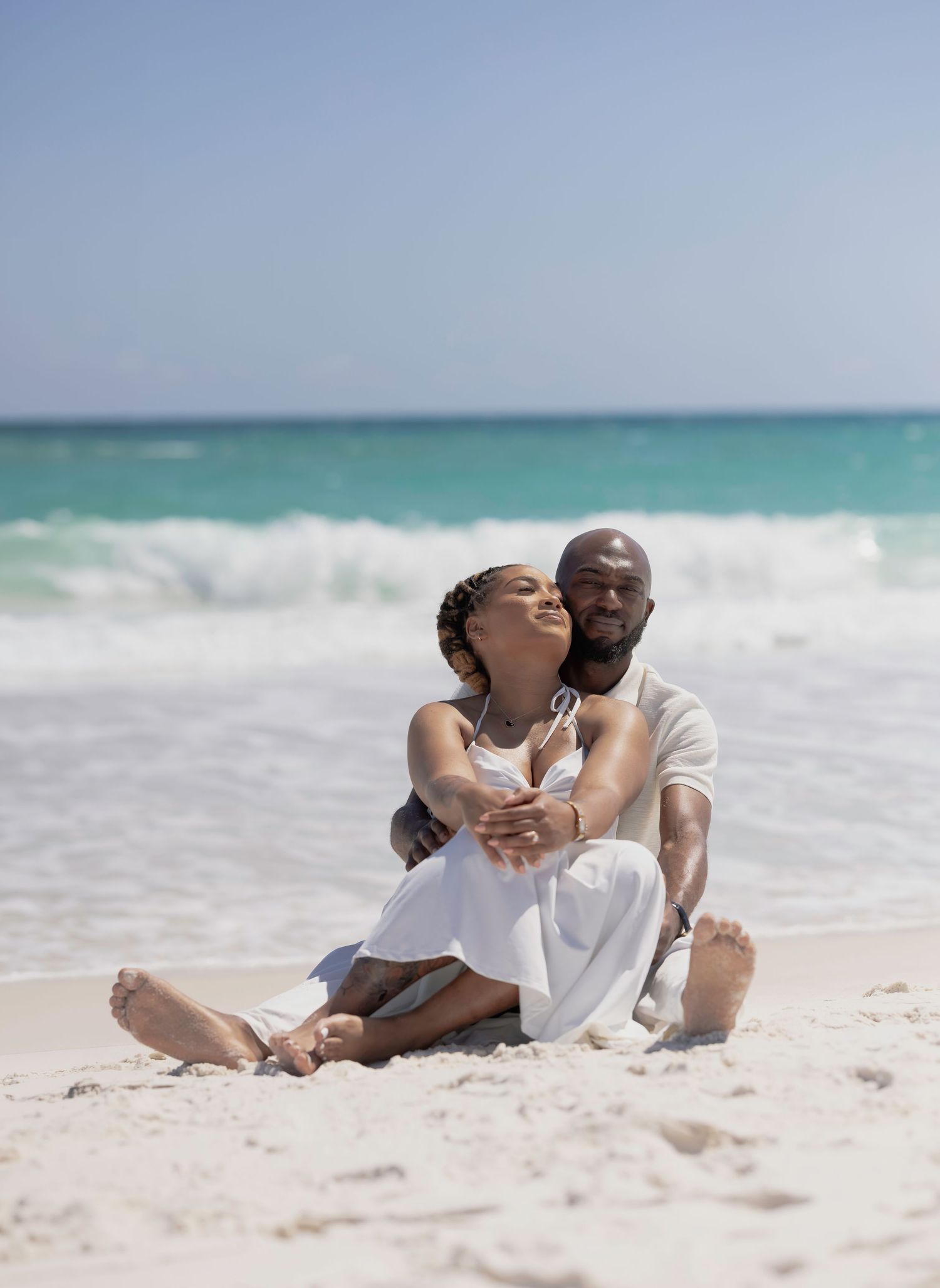 A couple in white sits together on sandy beach, embracing with turquoise ocean waves behind them.