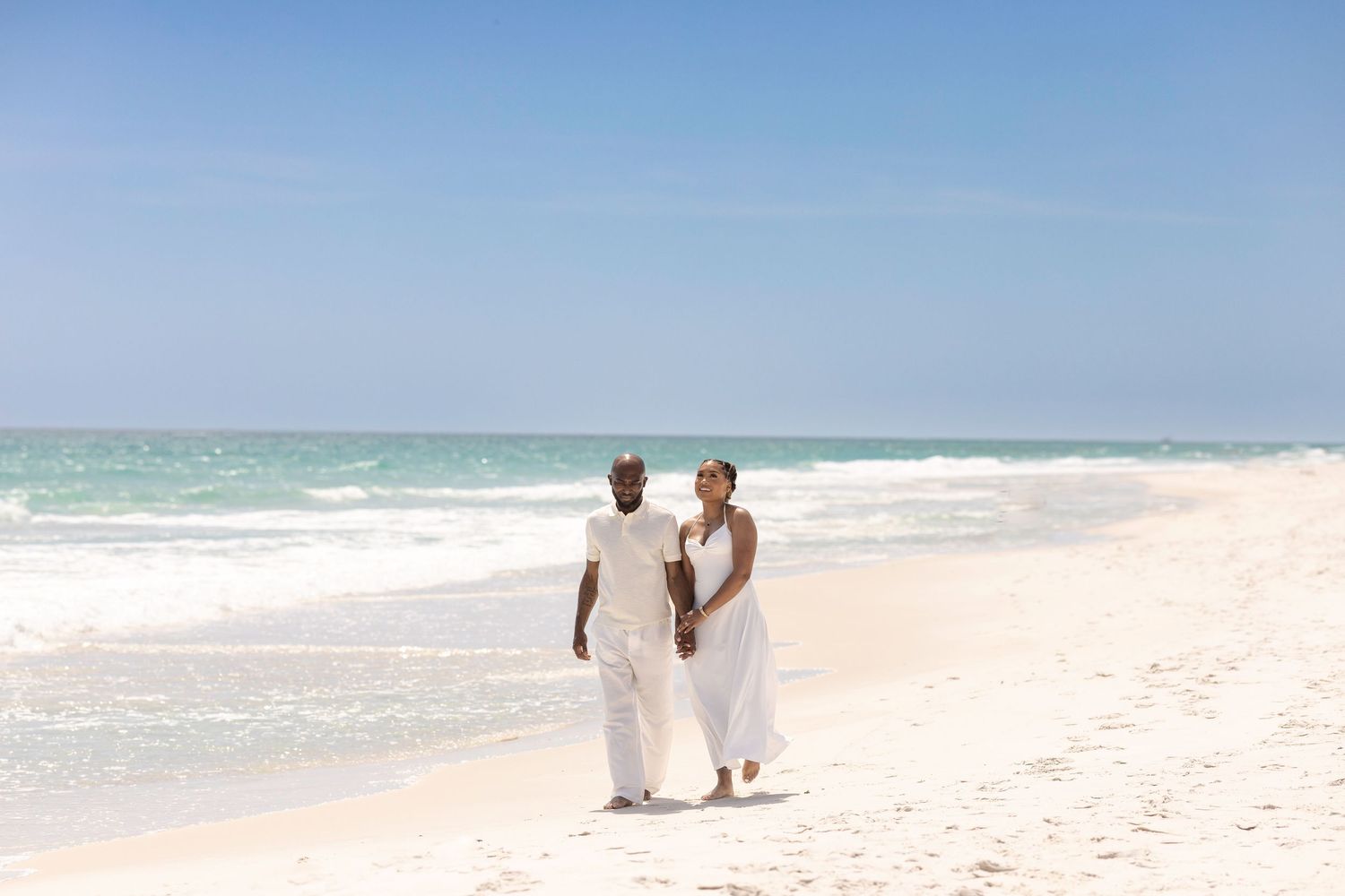 Couple in white attire walking barefoot on a pristine sandy beach with turquoise ocean waves in the background.