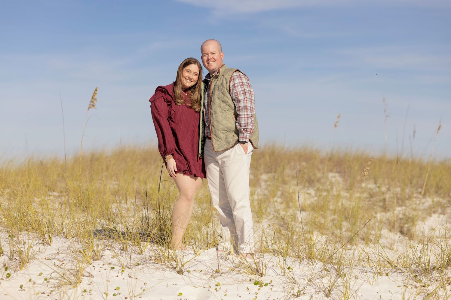 A smiling couple poses together on sandy beach dunes with sea oats on a sunny day.
