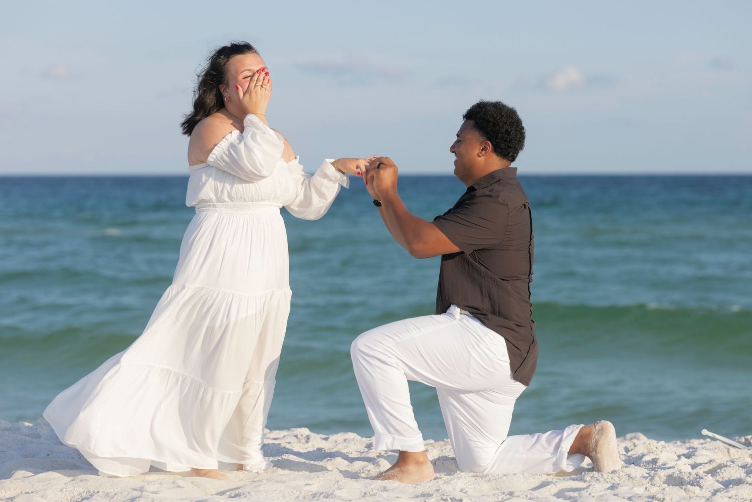 Man proposing on one knee to woman in white dress on a sunny beach, romantic marriage proposal scene.