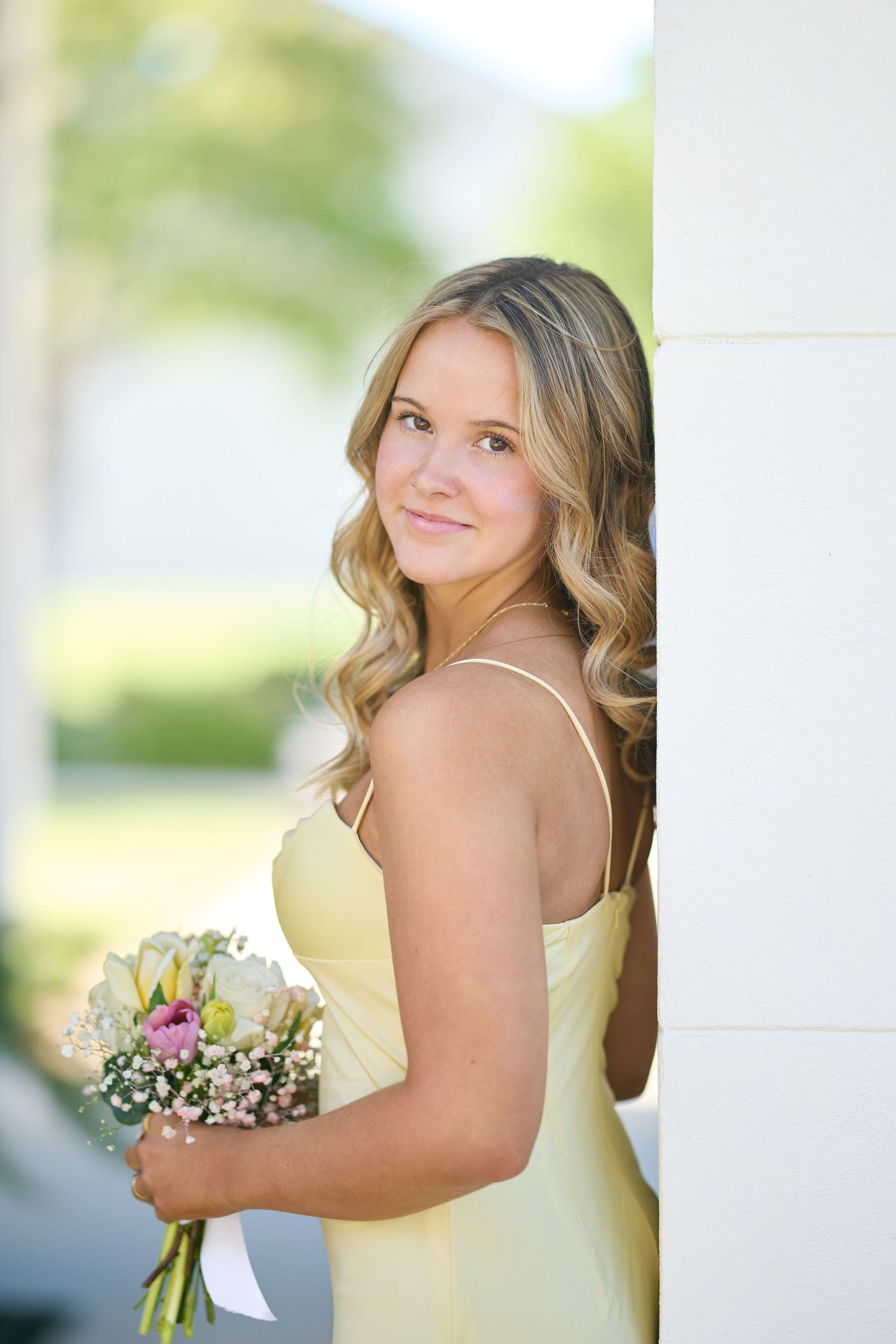 Young woman in yellow dress holding floral bouquet leans against white column outdoors.