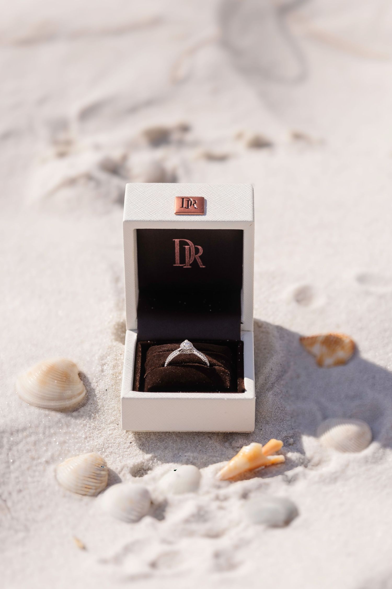 Diamond engagement ring in a DR jewelry box on white sand beach surrounded by seashells.