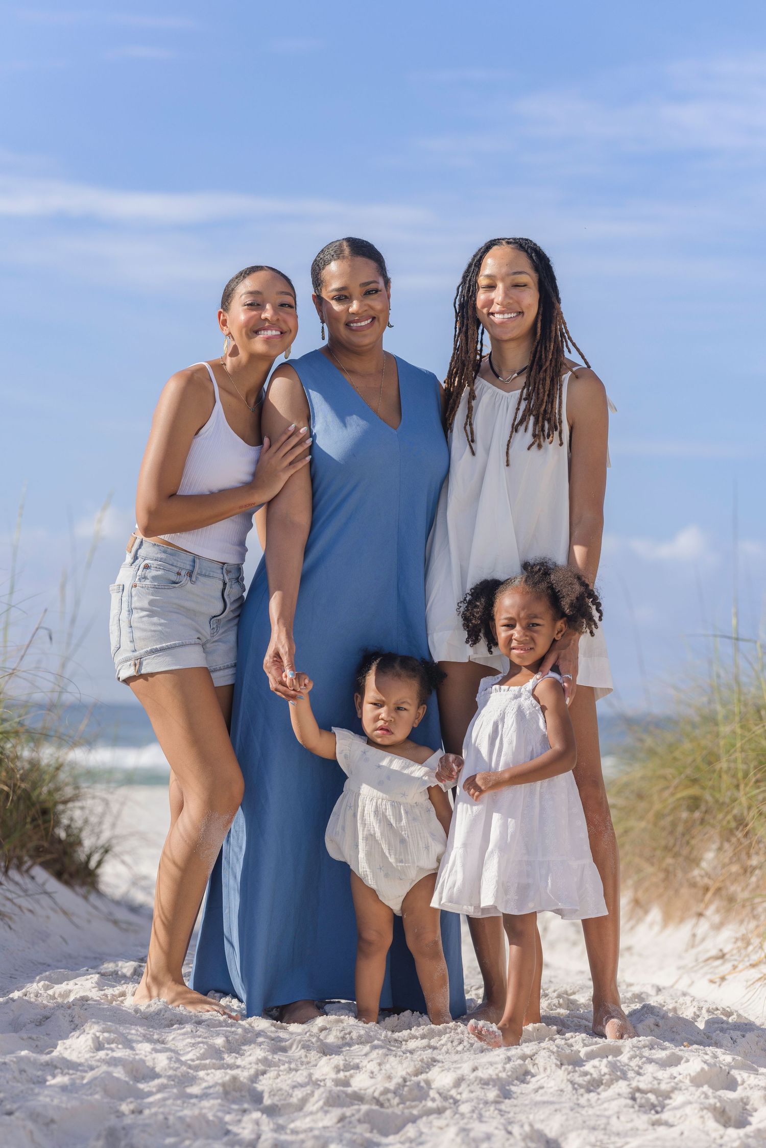 A joyful family of five poses on a sunny beach, with a mother in a blue dress surrounded by her four children.