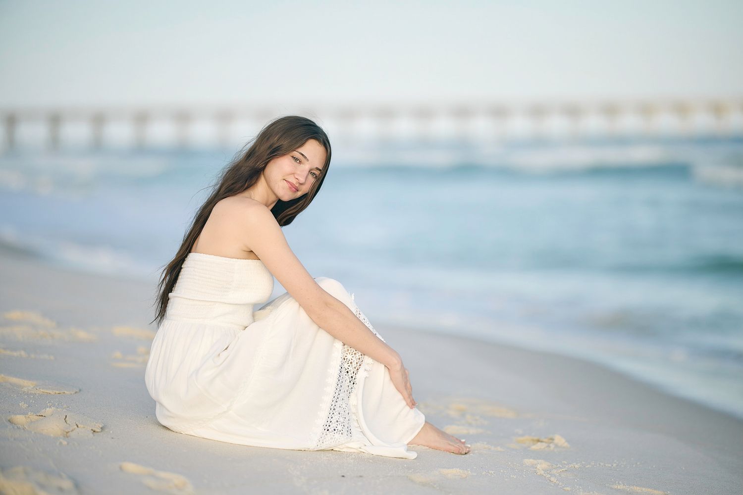 A person in a white dress sits on a sandy beach with a pier and ocean waves in the background.