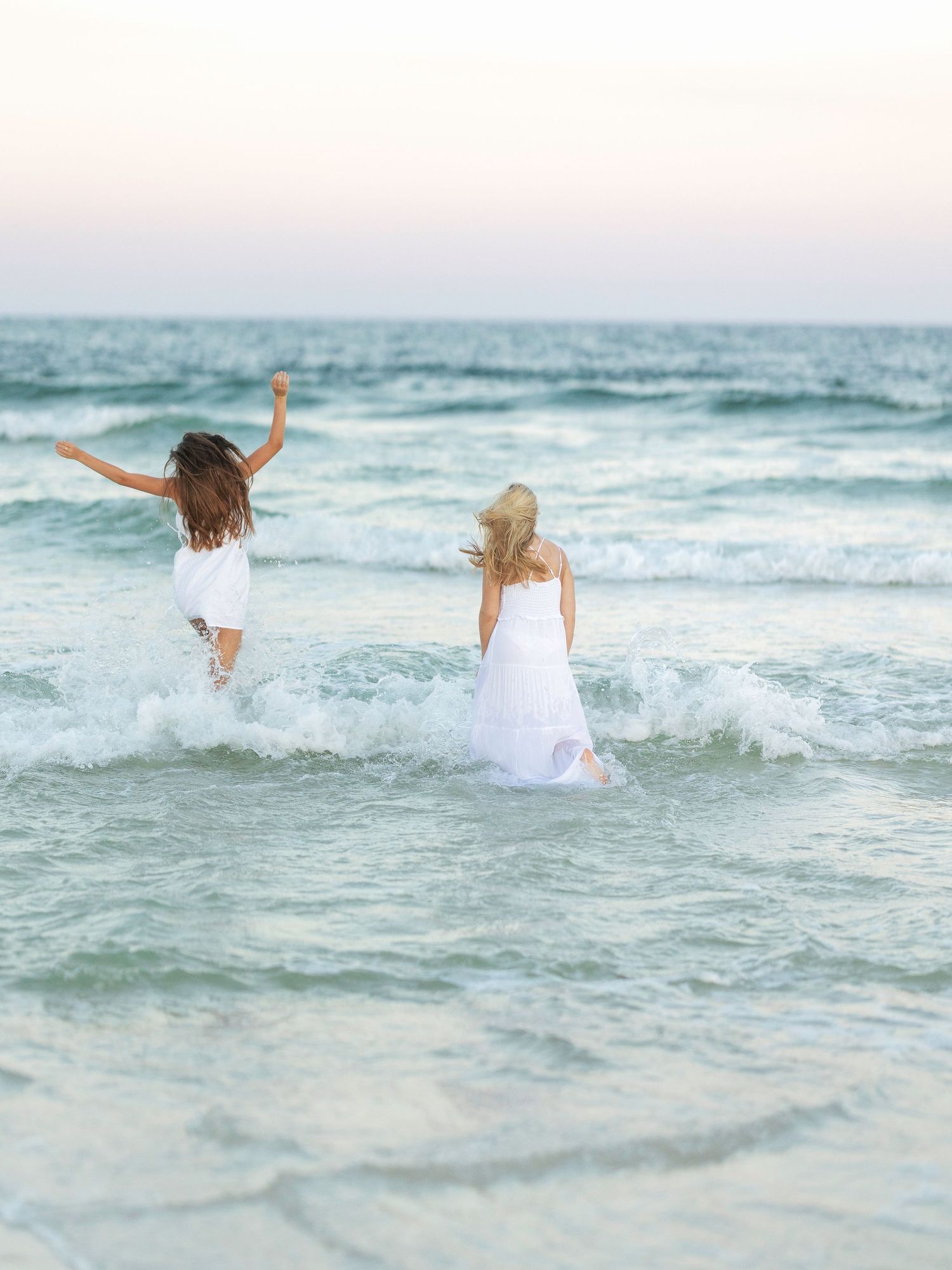 Two people in white dresses jumping and playing in ocean waves.