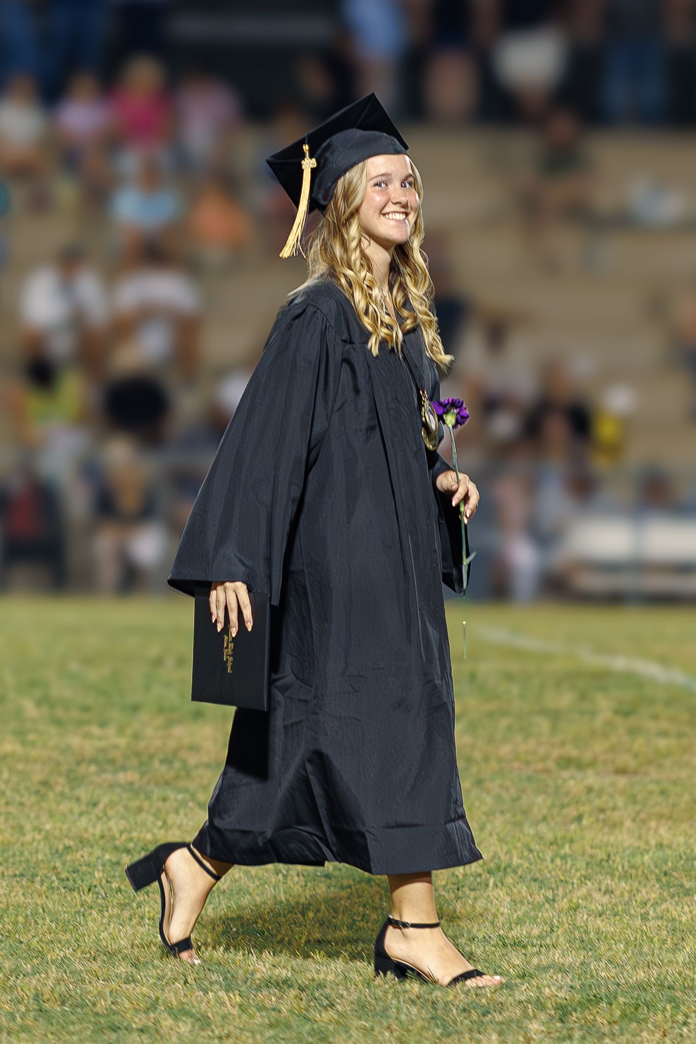Graduate smiles while walking across field in cap and gown holding purple flower.