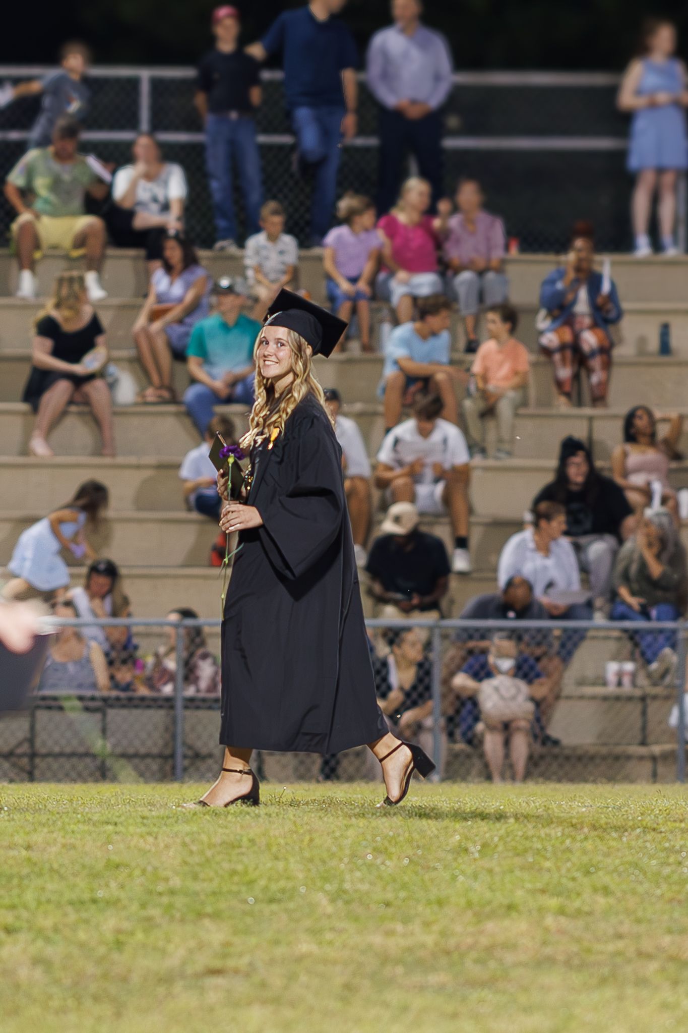 Graduate in black cap and gown walks across field during outdoor ceremony with spectators in bleachers.