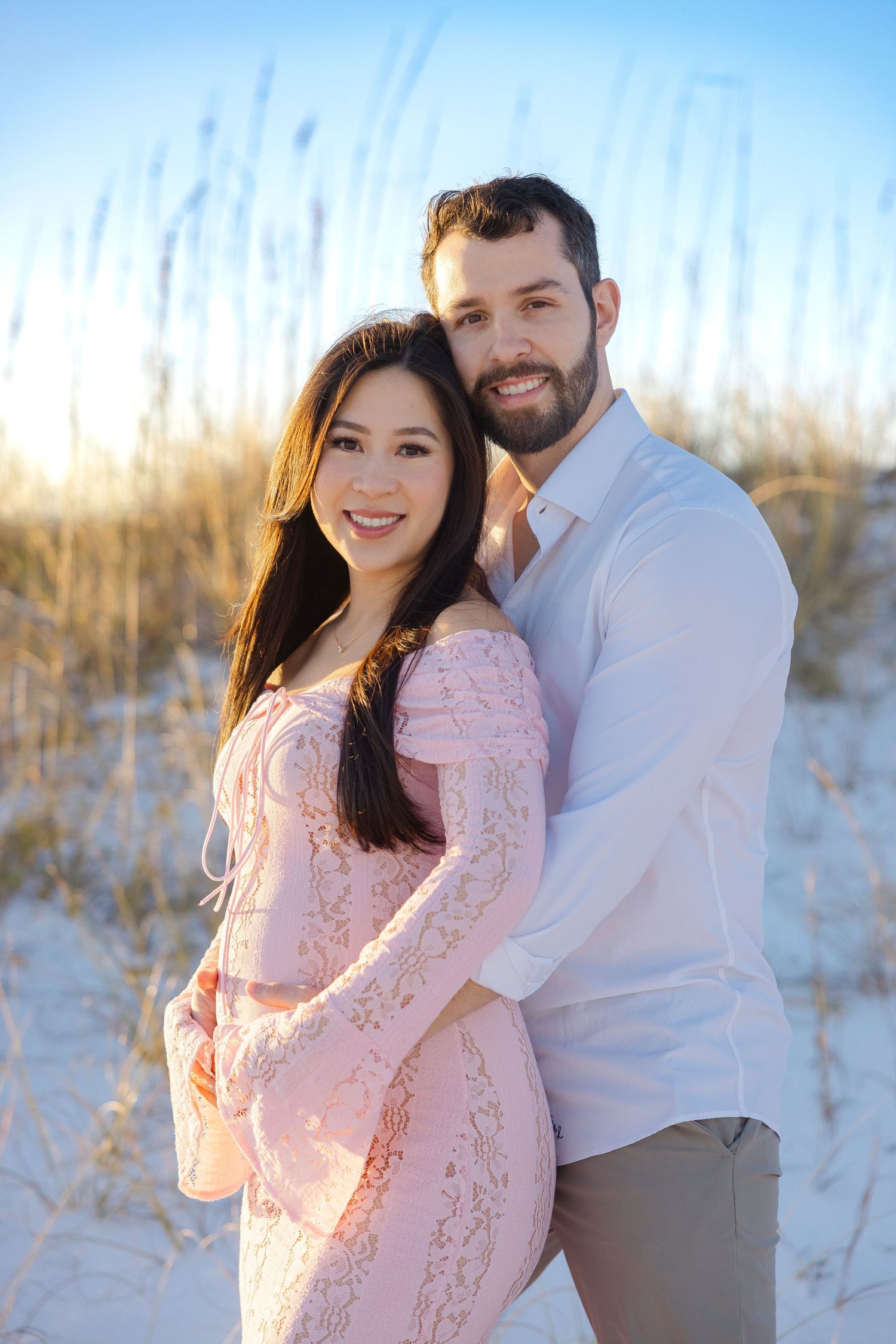 A couple embraces on a sandy beach with sea grass and dunes during golden hour in a romantic photoshoot.