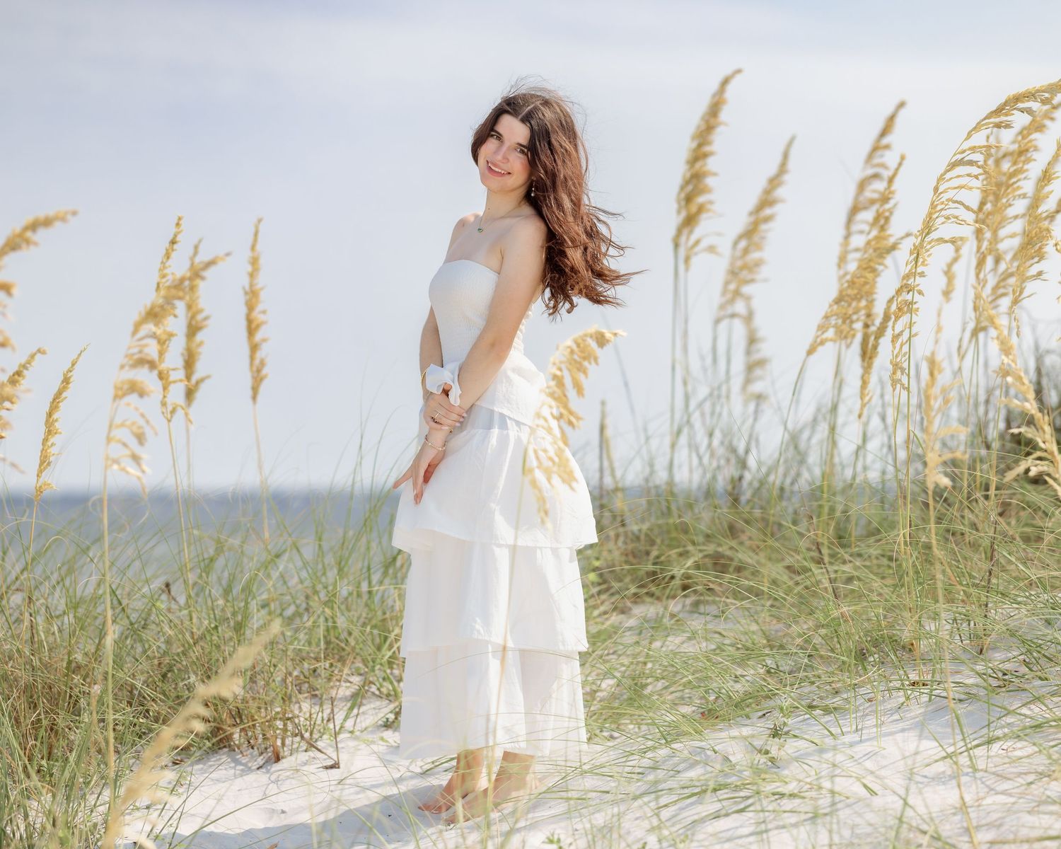 Brunette woman in white layered dress standing among tall sea oats on beach dunes.