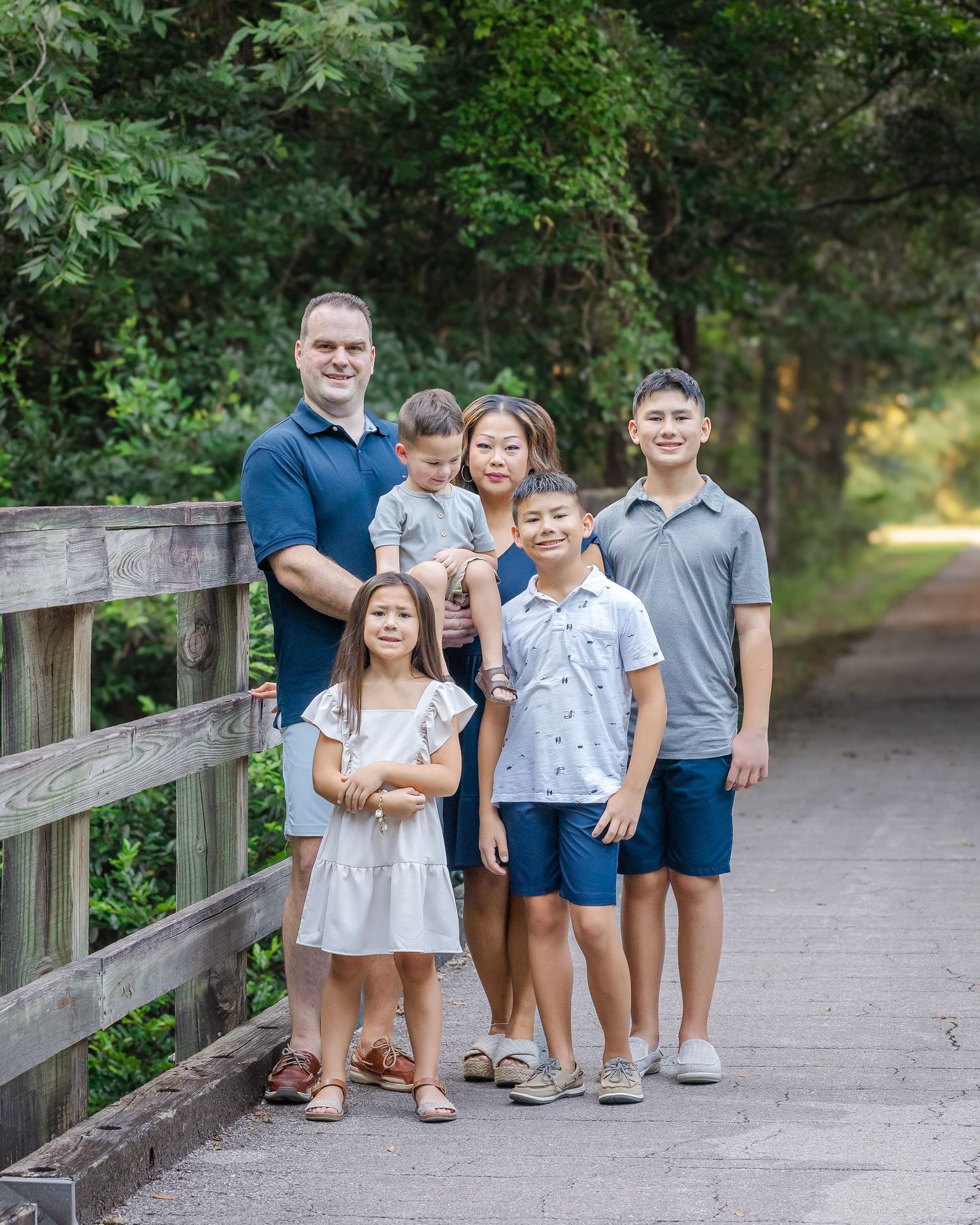Family of six posing on a wooden bridge path surrounded by lush green trees during an outdoor photo session.