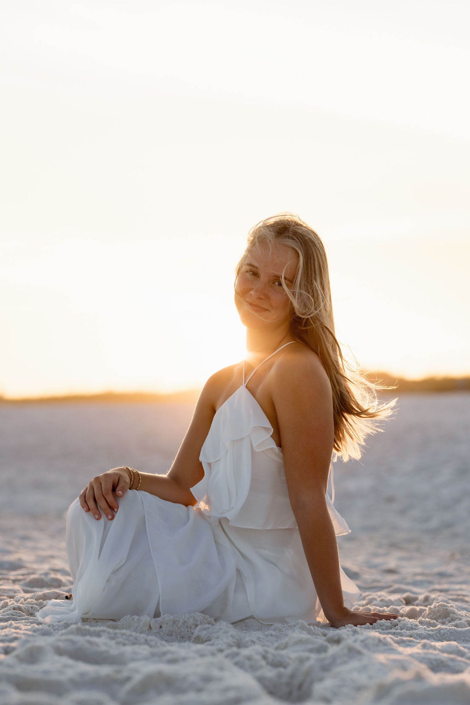 Young blonde woman in white dress sitting on sandy beach at golden sunset, smiling over shoulder.