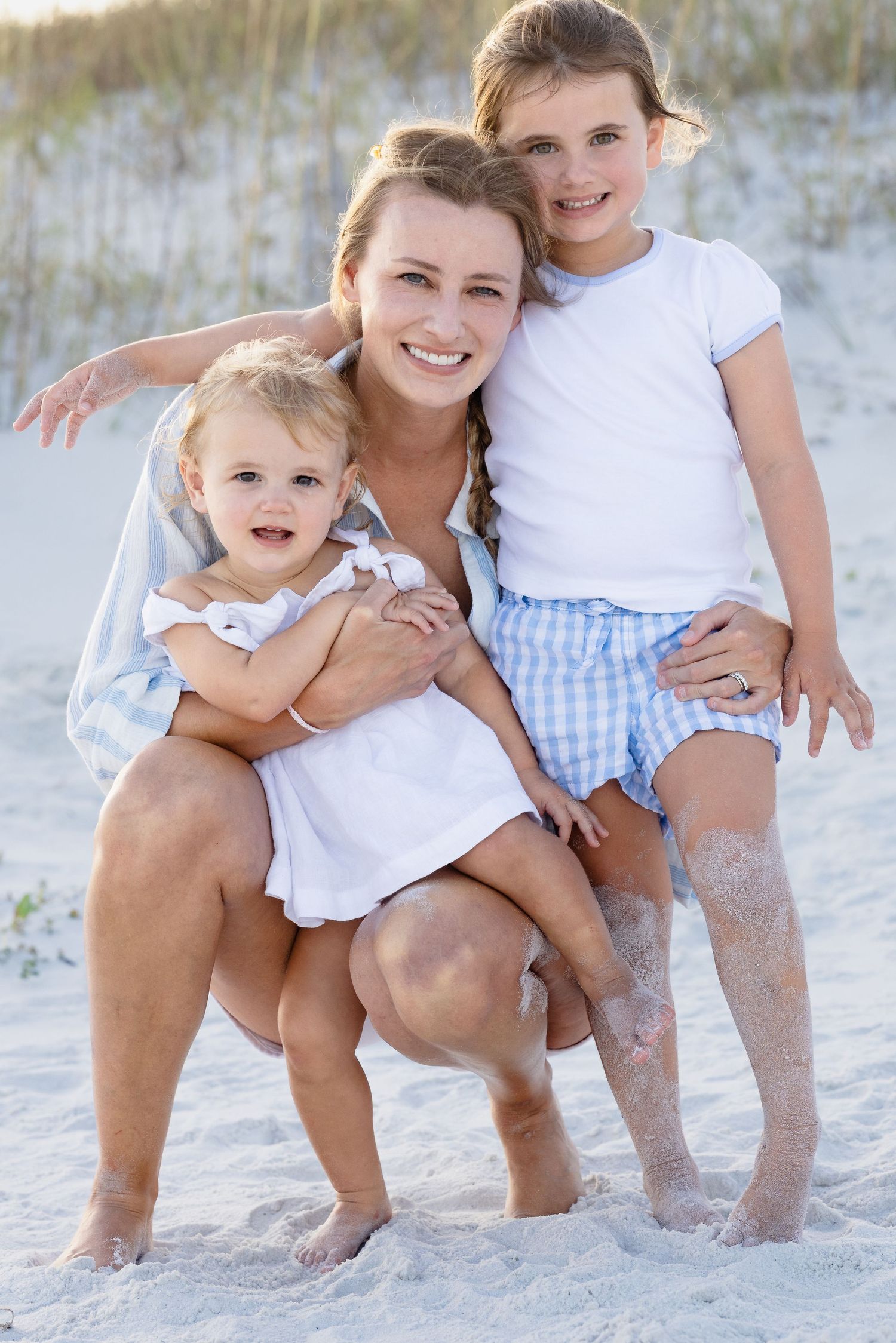 A mother crouching on a sandy beach, smiling while hugging her two young daughters.