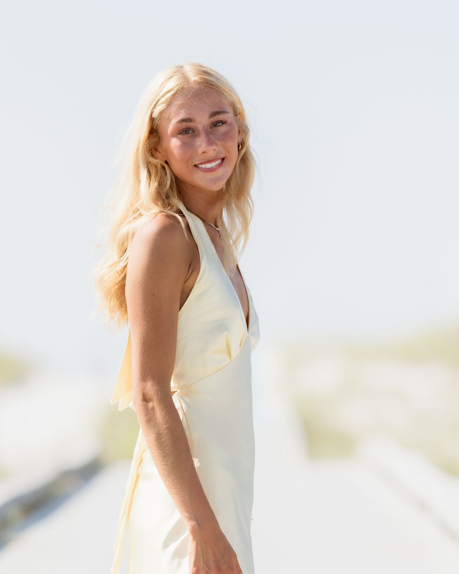 Blonde woman in cream halter dress smiling over shoulder on sunny white sand beach.