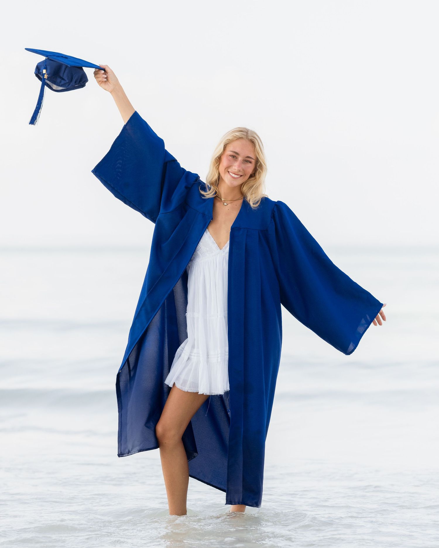 Smiling blonde graduate in royal blue cap and gown standing in ocean waves wearing white dress.