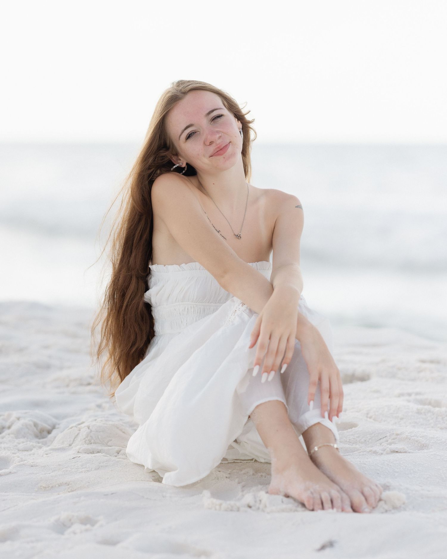Young woman in white strapless dress sitting barefoot on sandy beach with ocean in background.