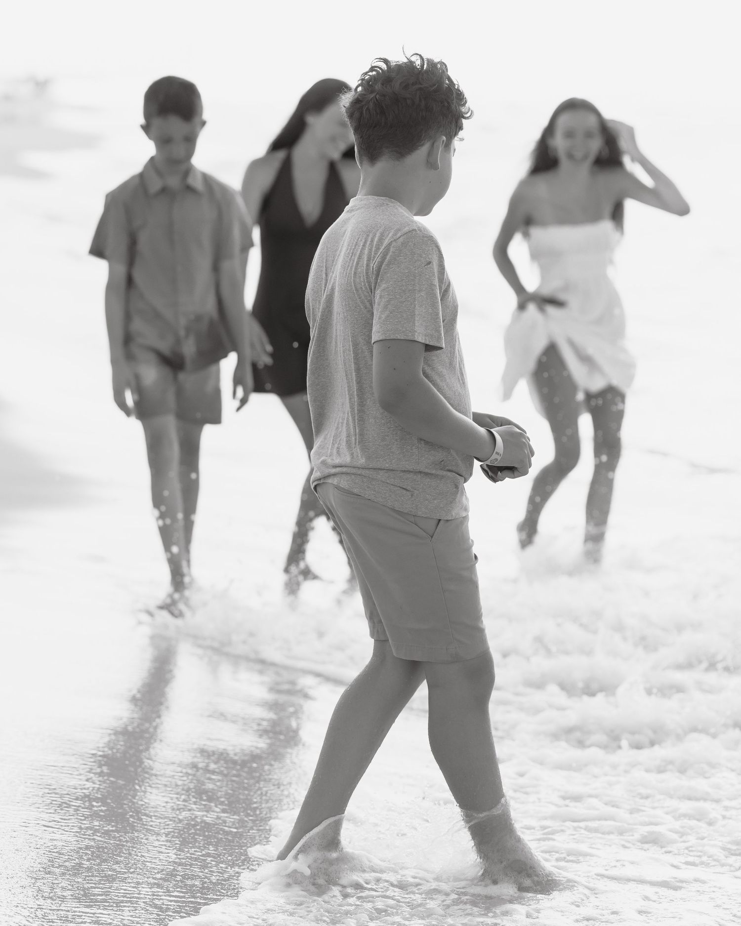 Black and white photo of four people walking on a beach in shallow ocean waves.