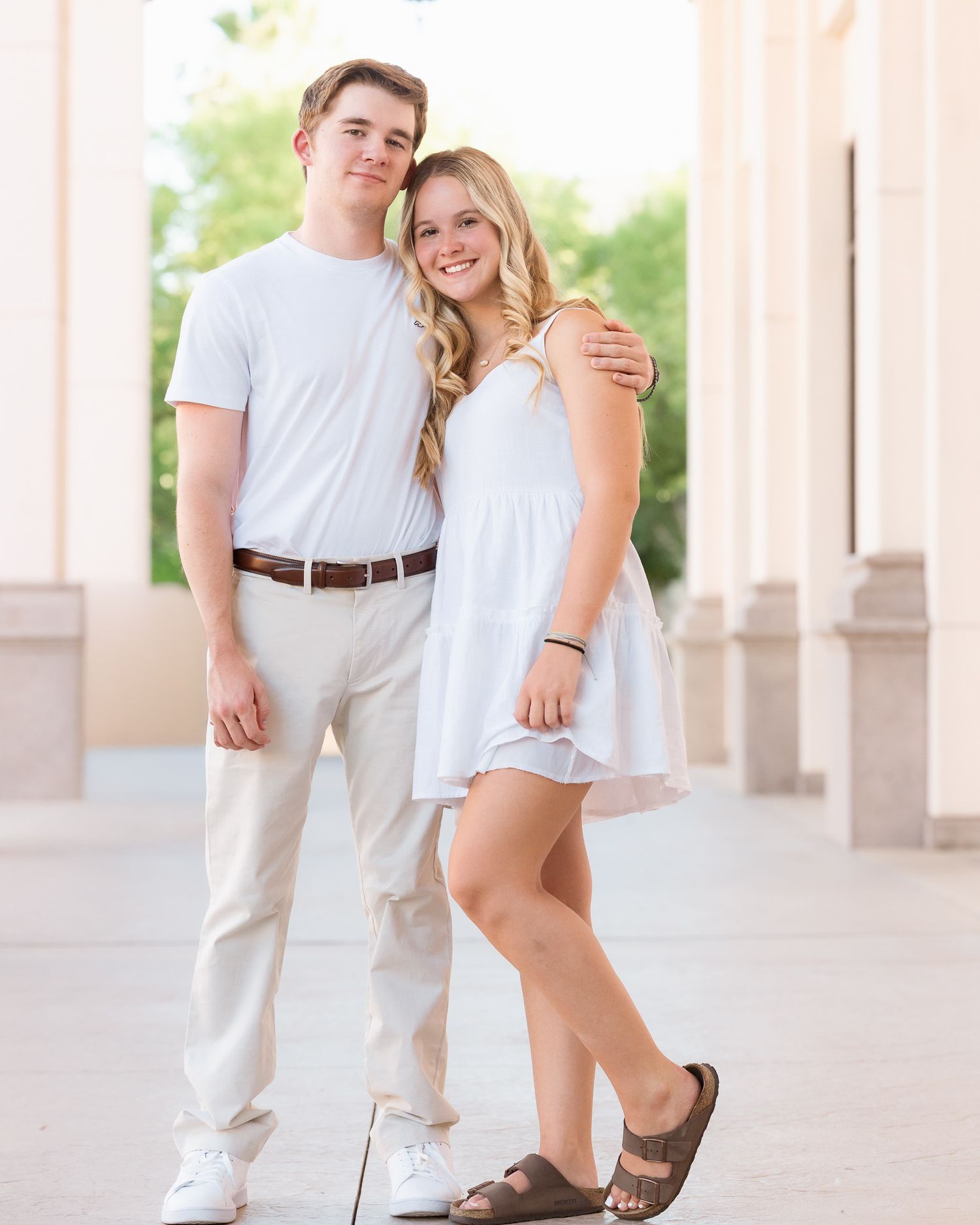 A young couple in matching white outfits posing together outdoors near elegant columns.