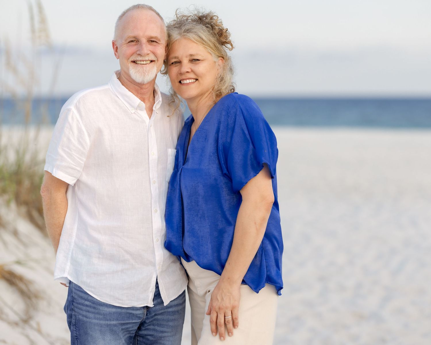 Happy senior couple embracing on a sandy beach, man in white shirt and woman in blue top smiling.