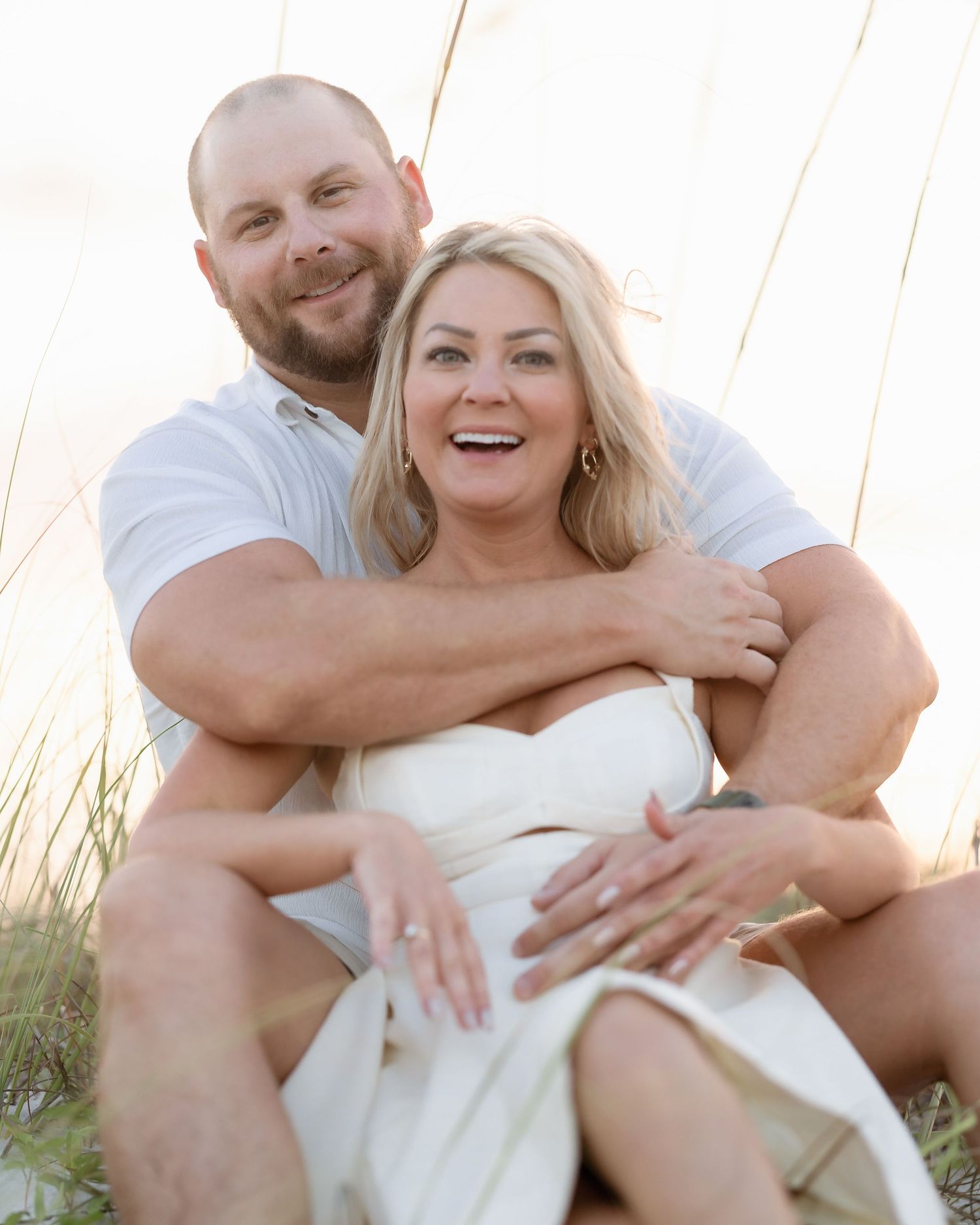 A smiling couple sits together in beach grass, the man embracing the woman from behind, both wearing white outfits.