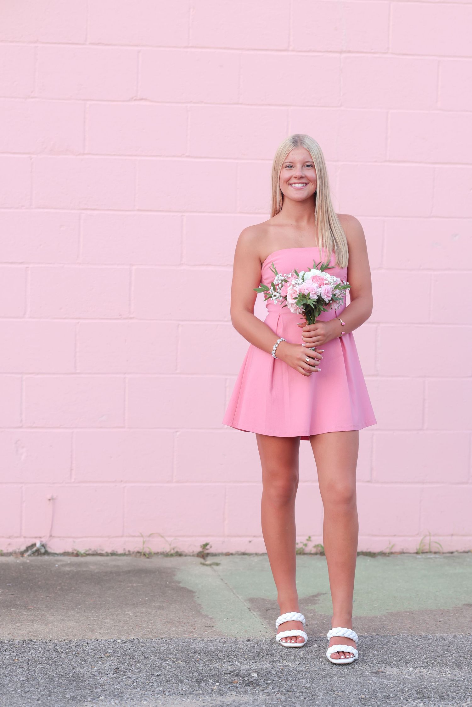 A person in a pink dress stands against a pink wall, their lower half shown holding white flowers and wearing white sandals.