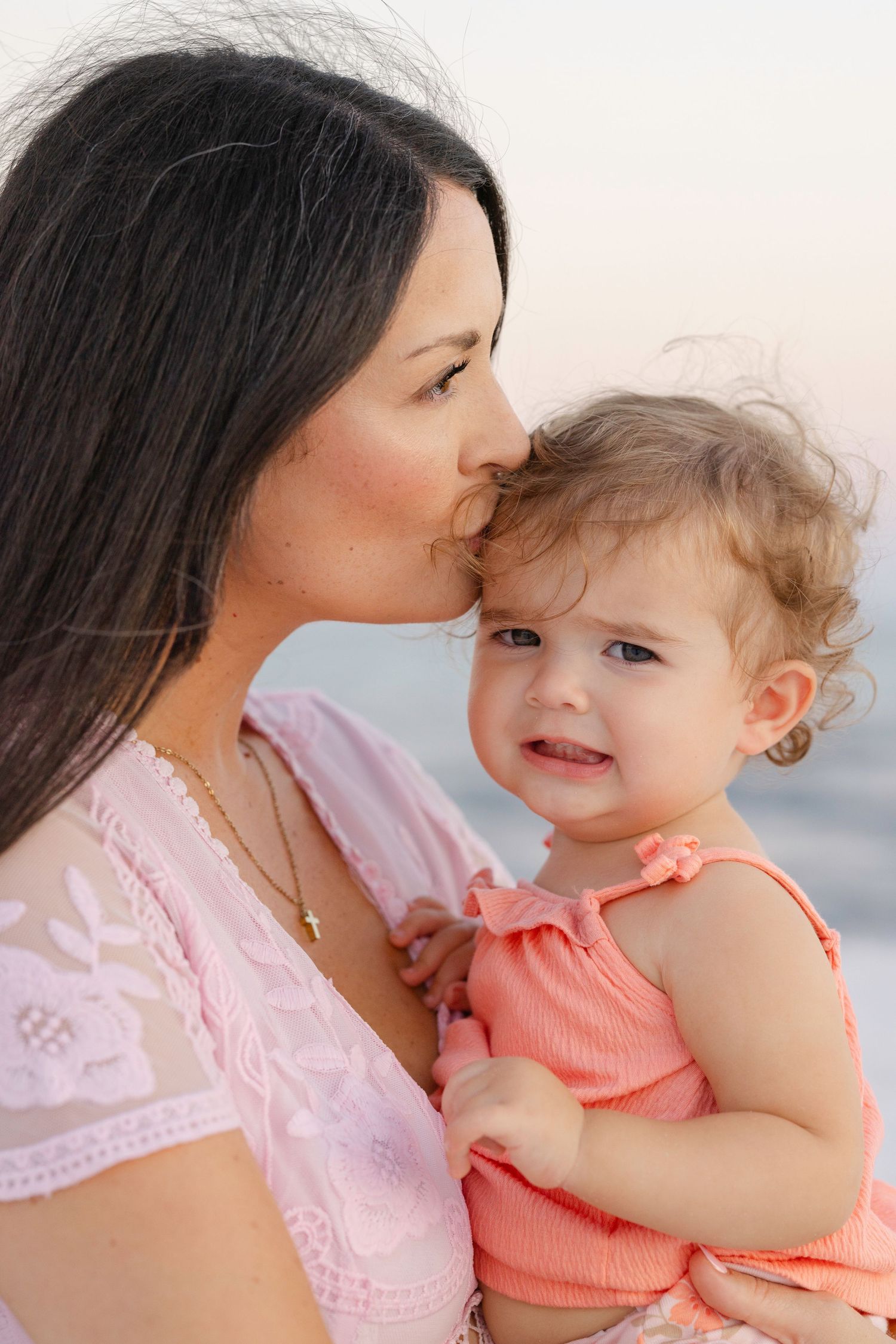 A tender moment between parent and baby against an ocean backdrop while wearing pink and coral outfits.